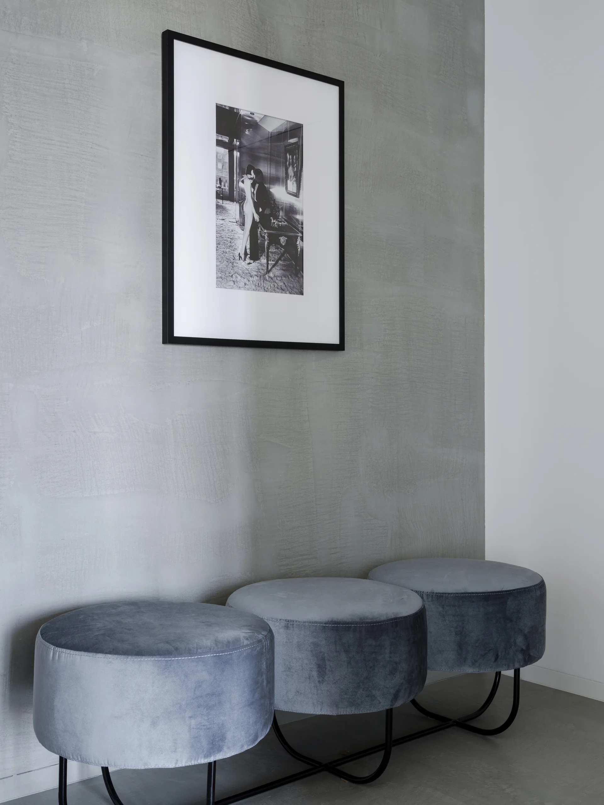 Three round grey velvet ottomans stand in a line against a textured grey plaster wall underneath a framed black and white picture.