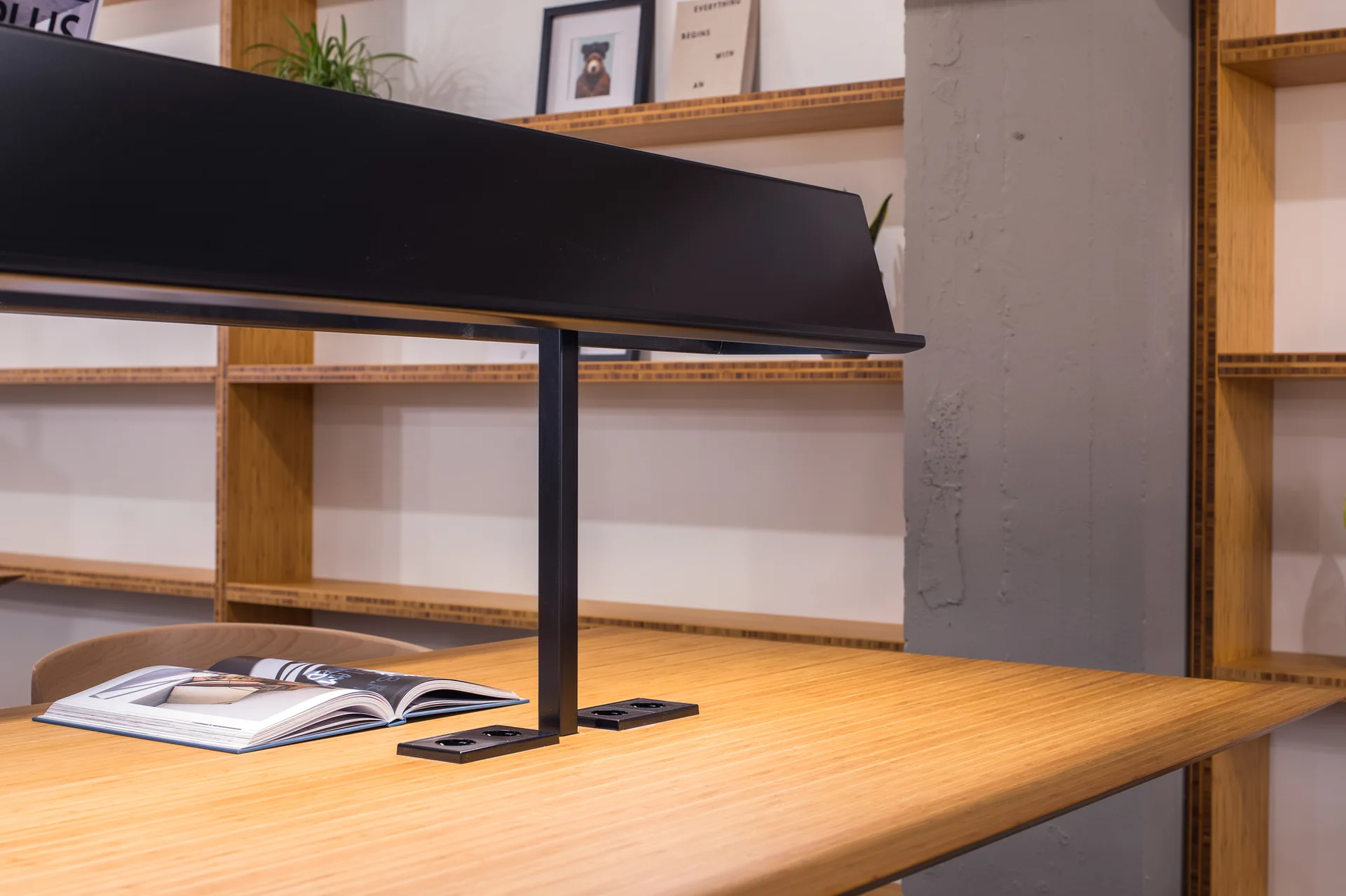 A warm wood library desk features an integrated black metal task lamp with open bookshelves and a concrete pillar in the background.
