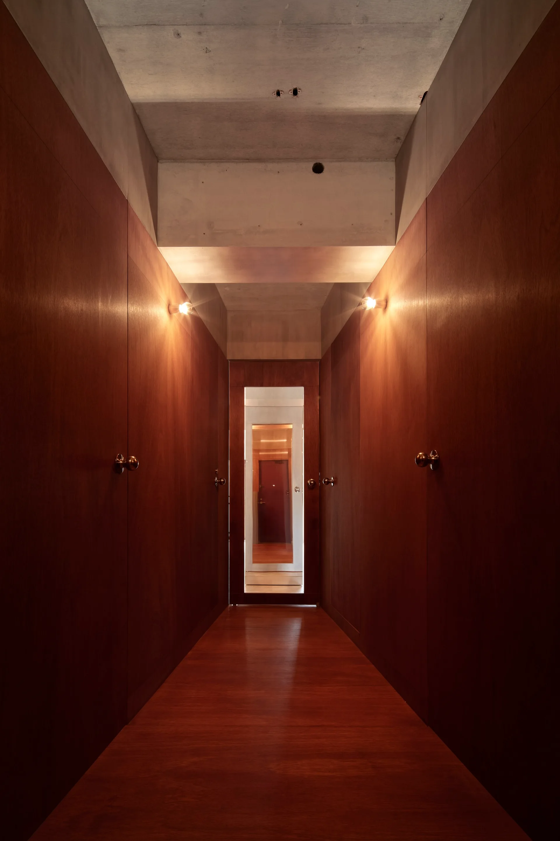 A narrow residential hallway featuring dark wood paneling, matching wood floors, warm wall-mounted sconces, and an exposed concrete ceiling.