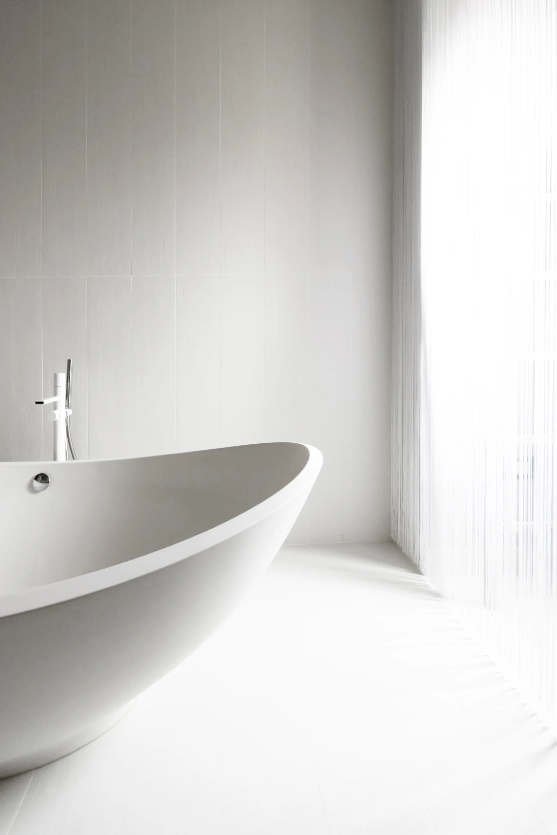 A minimalist white bathroom featuring a curved freestanding tub next to a window with sheer white curtains and subtle vertical wall paneling.