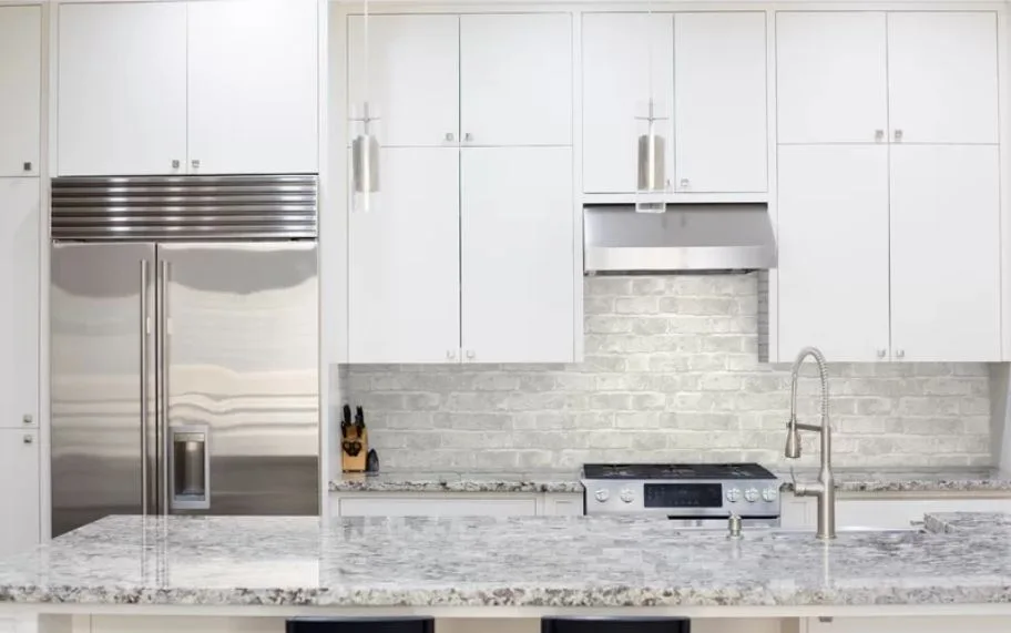 A modern kitchen features white flat-panel cabinetry, a stainless steel refrigerator, and a speckled grey stone island countertop.