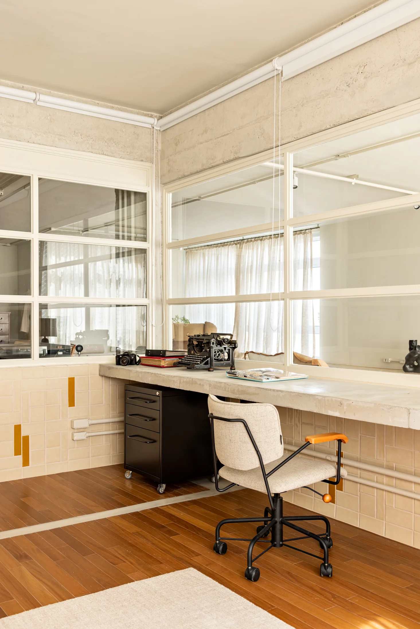 A home office features a floating concrete desk, a fabric office chair, a black rolling cabinet, and a glass partition wall.