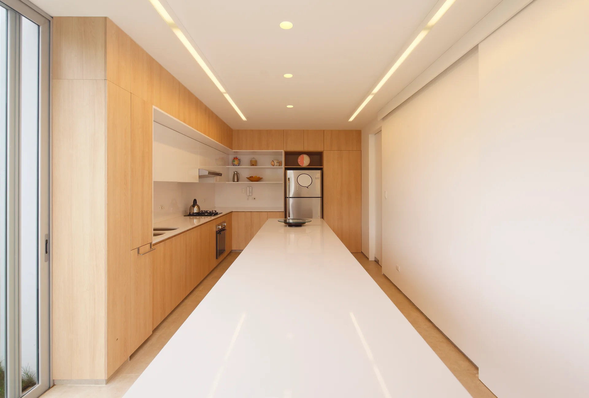 A minimalist kitchen featuring an extra long white island, light wood cabinets, built-in appliances, and linear ceiling lighting.