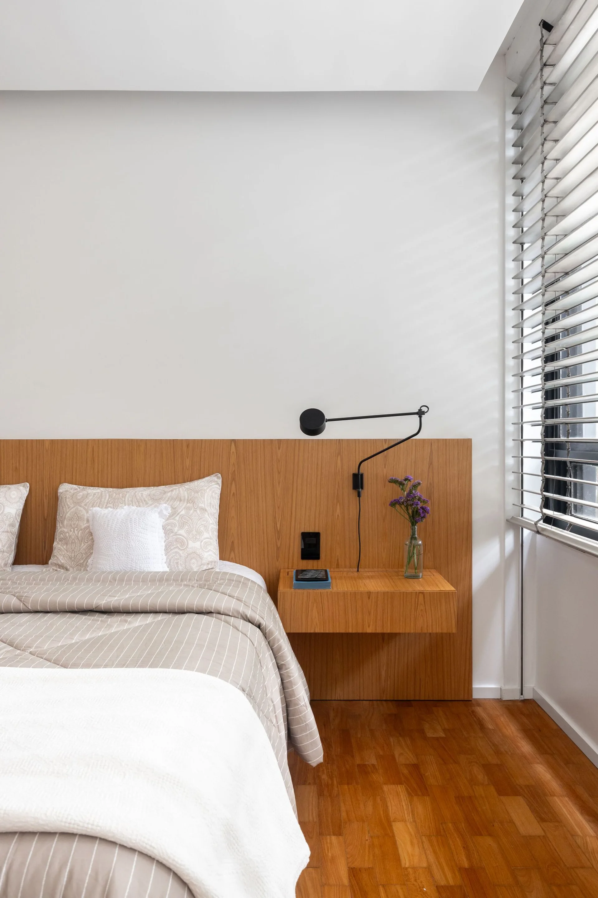 A modern bedroom features a wide wooden headboard panel with an integrated floating nightstand, a black wall sconce, and striped bedding.