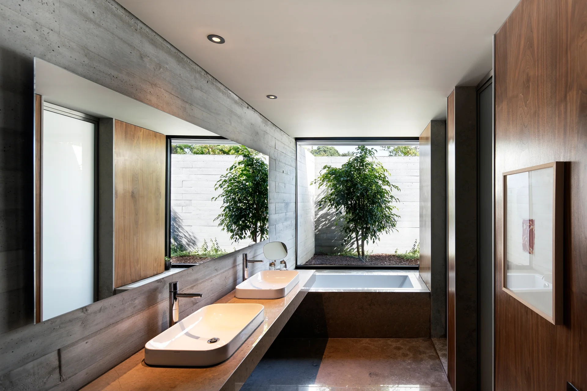 A minimalist bathroom with board-formed concrete walls, a long wood vanity, two white vessel sinks, and a large window looking out onto a small tree.
