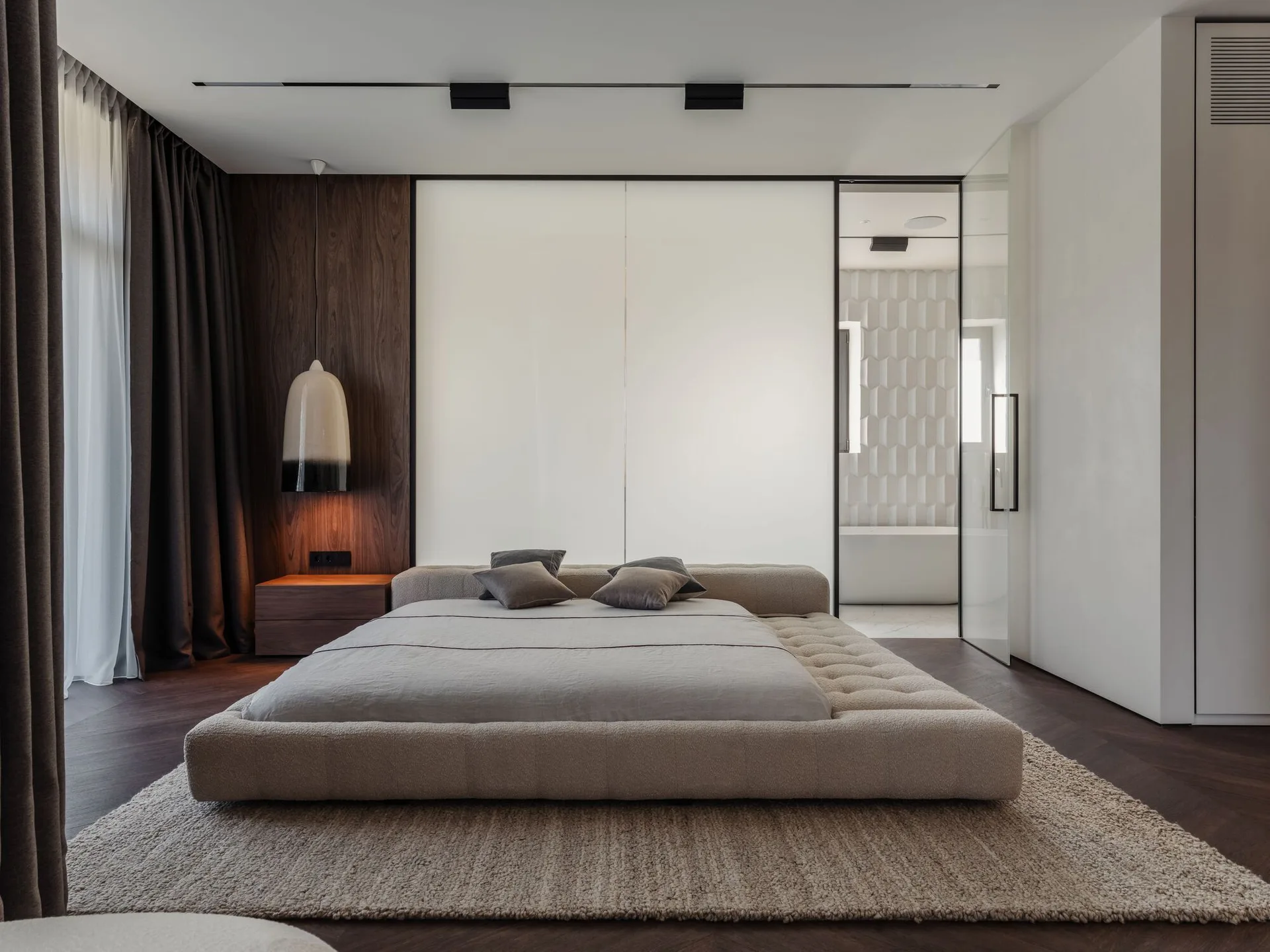 A low-profile upholstered platform bed sits on a shag rug in a modern bedroom featuring dark wood paneling and a frosted glass partition wall.