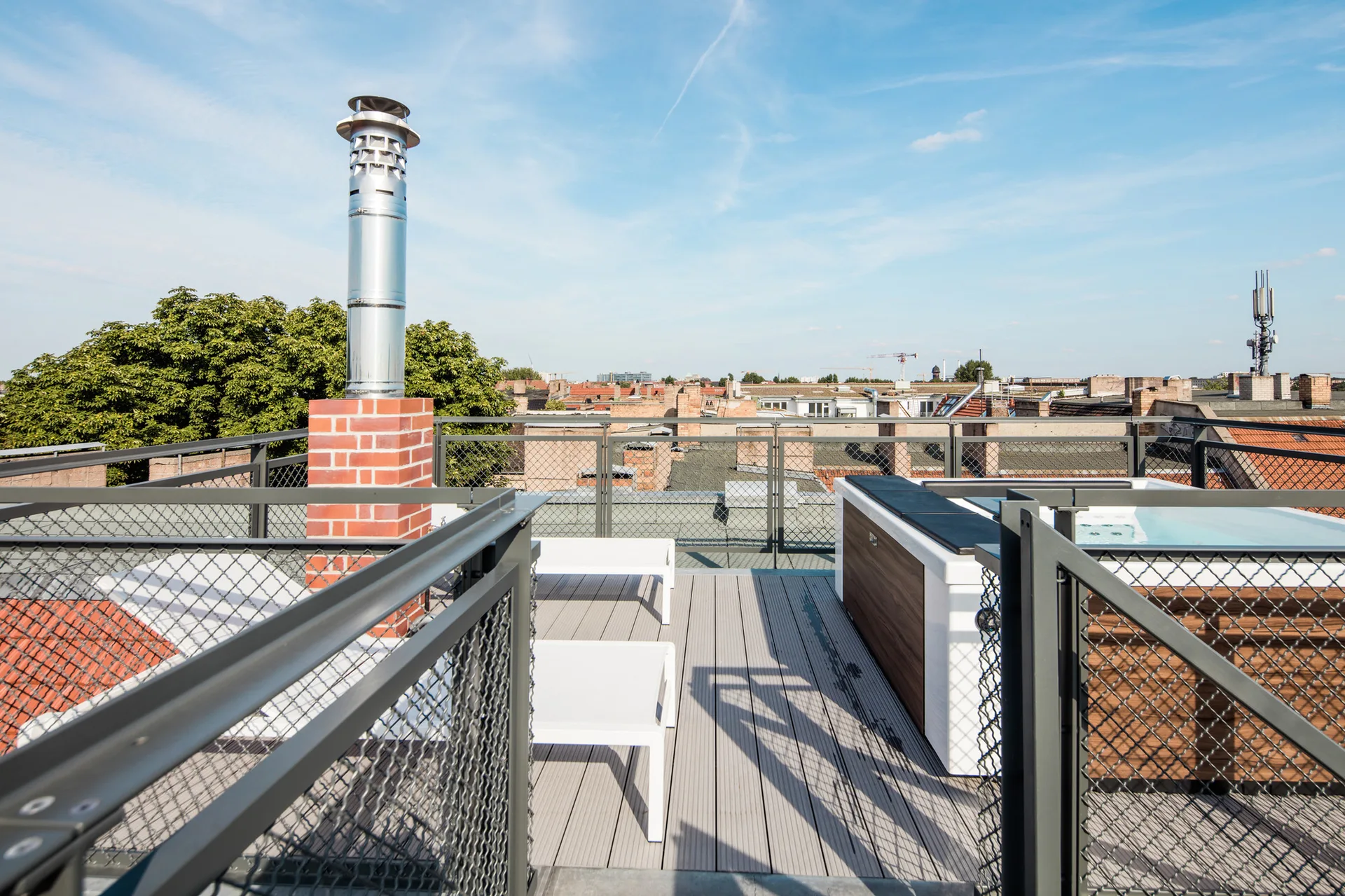 A modern outdoor rooftop terrace features grey wood-look decking, a white hot tub with wood paneling, white lounge chairs, and metal mesh railings.