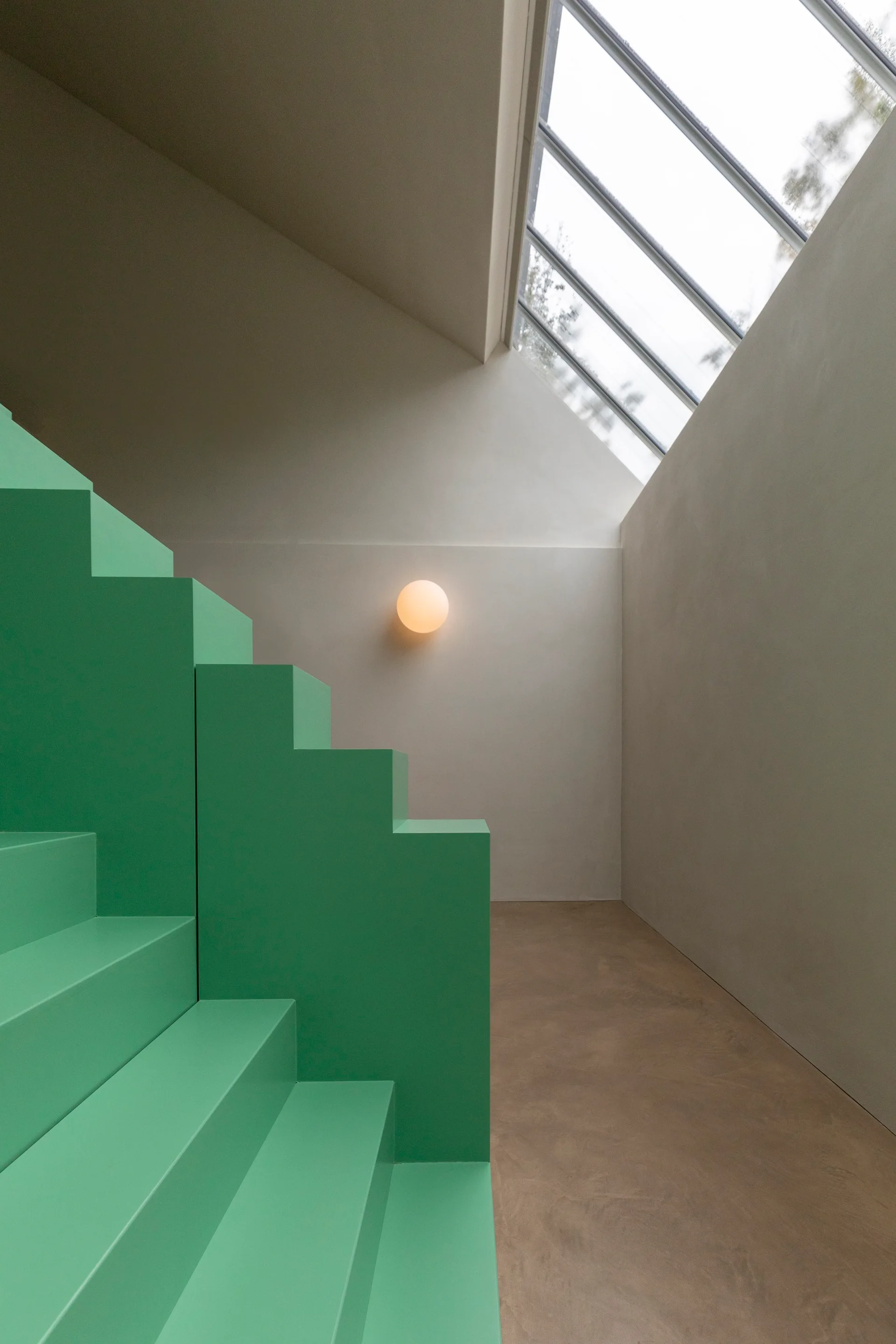 A minimalist stairwell features a bold mint green geometric staircase, a slanted skylight overhead, and a glowing round sconce on a plain wall.