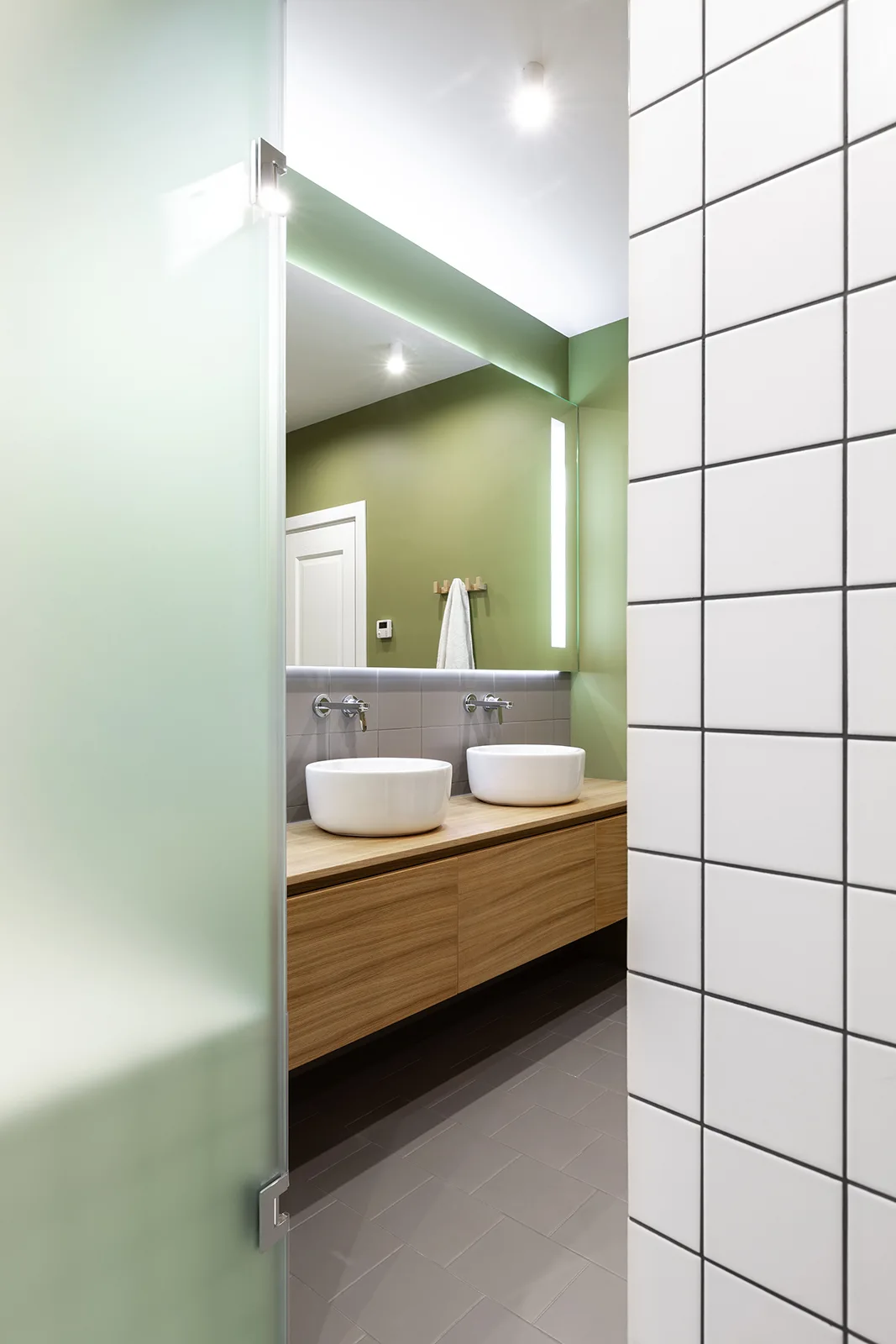 A contemporary bathroom with a floating wood vanity, white vessel sinks, a sage green wall, and white square tiled walls.