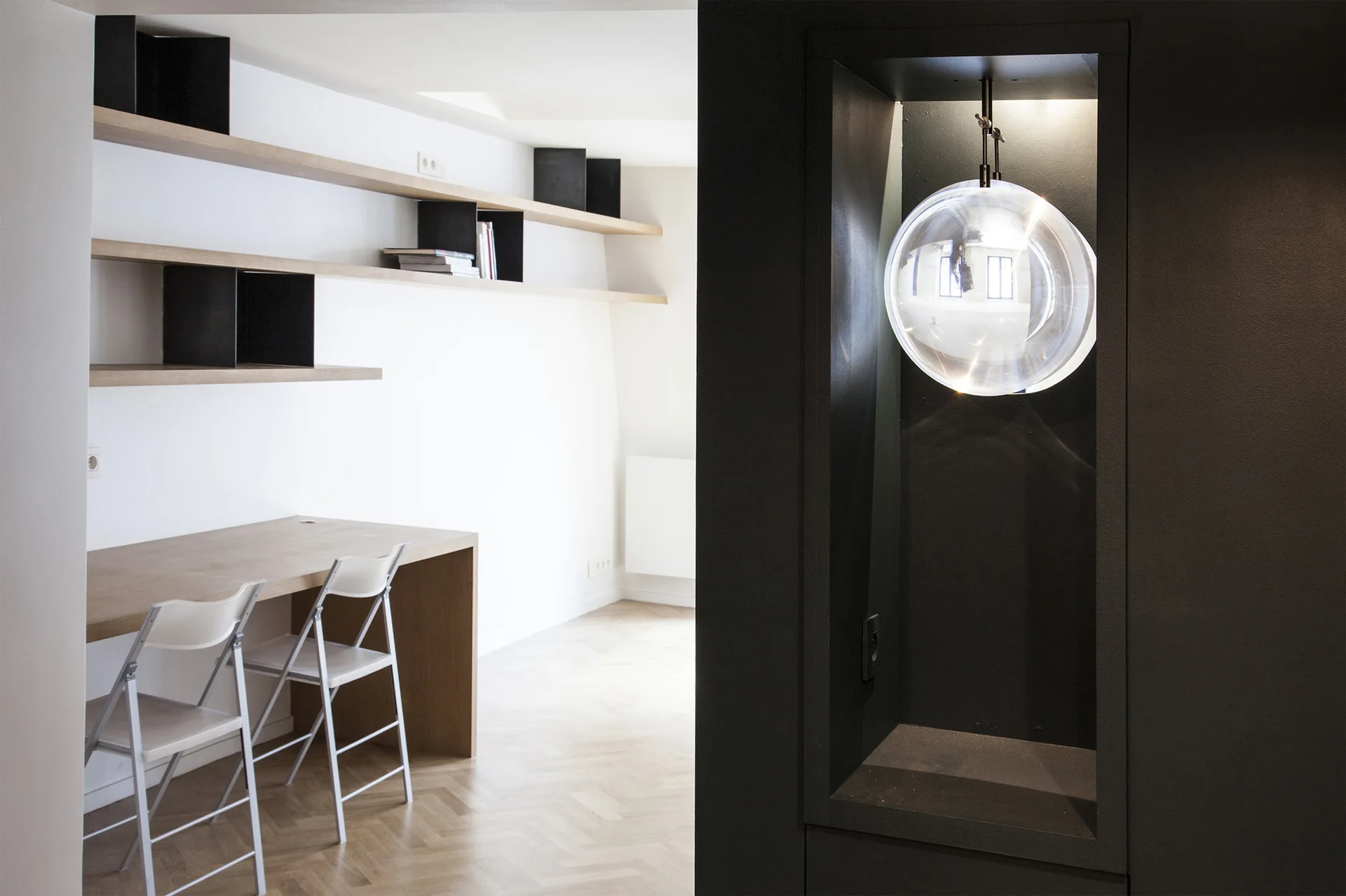 A minimalist home office featuring a light wood built-in desk, white folding chairs, floating wall shelves, and a dark alcove with a glass globe pendant light.