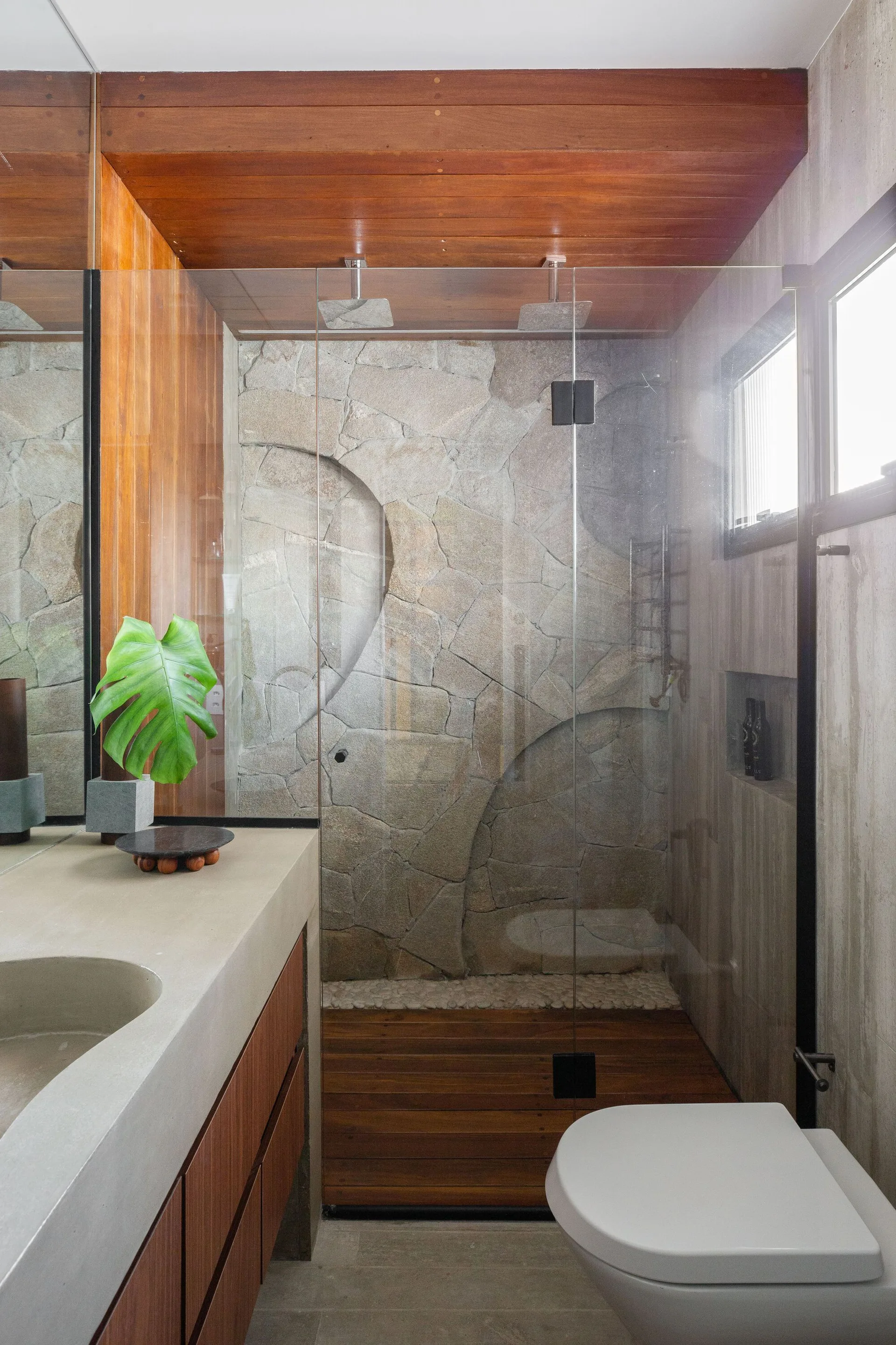 A contemporary bathroom features a walk-in glass shower with an irregular stone wall, a warm wood-plank ceiling, and a concrete vanity.