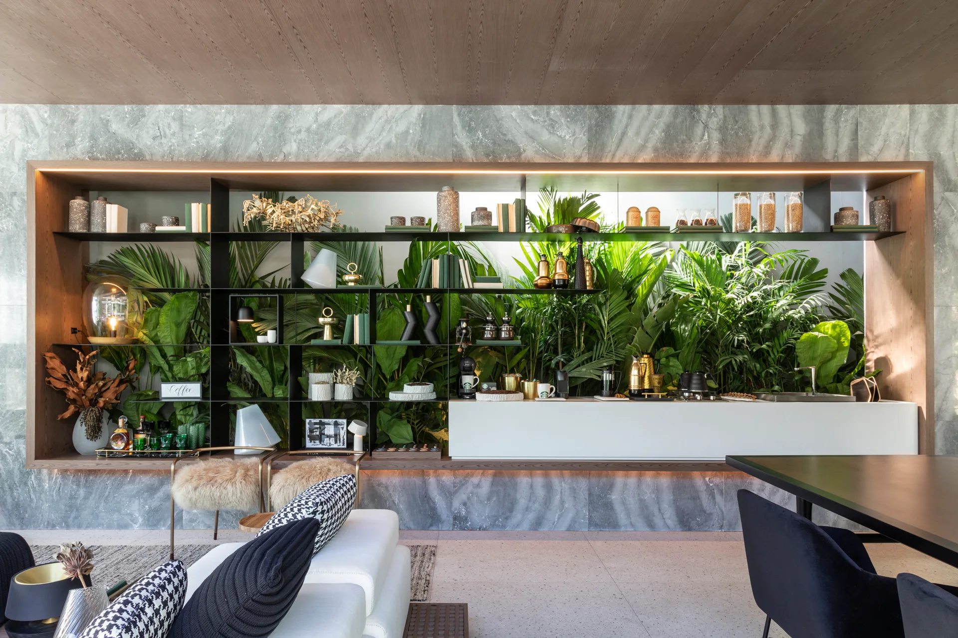 A large black metal shelving unit displaying decor stands against a lush green plant wall in a modern living space with grey stone walls.