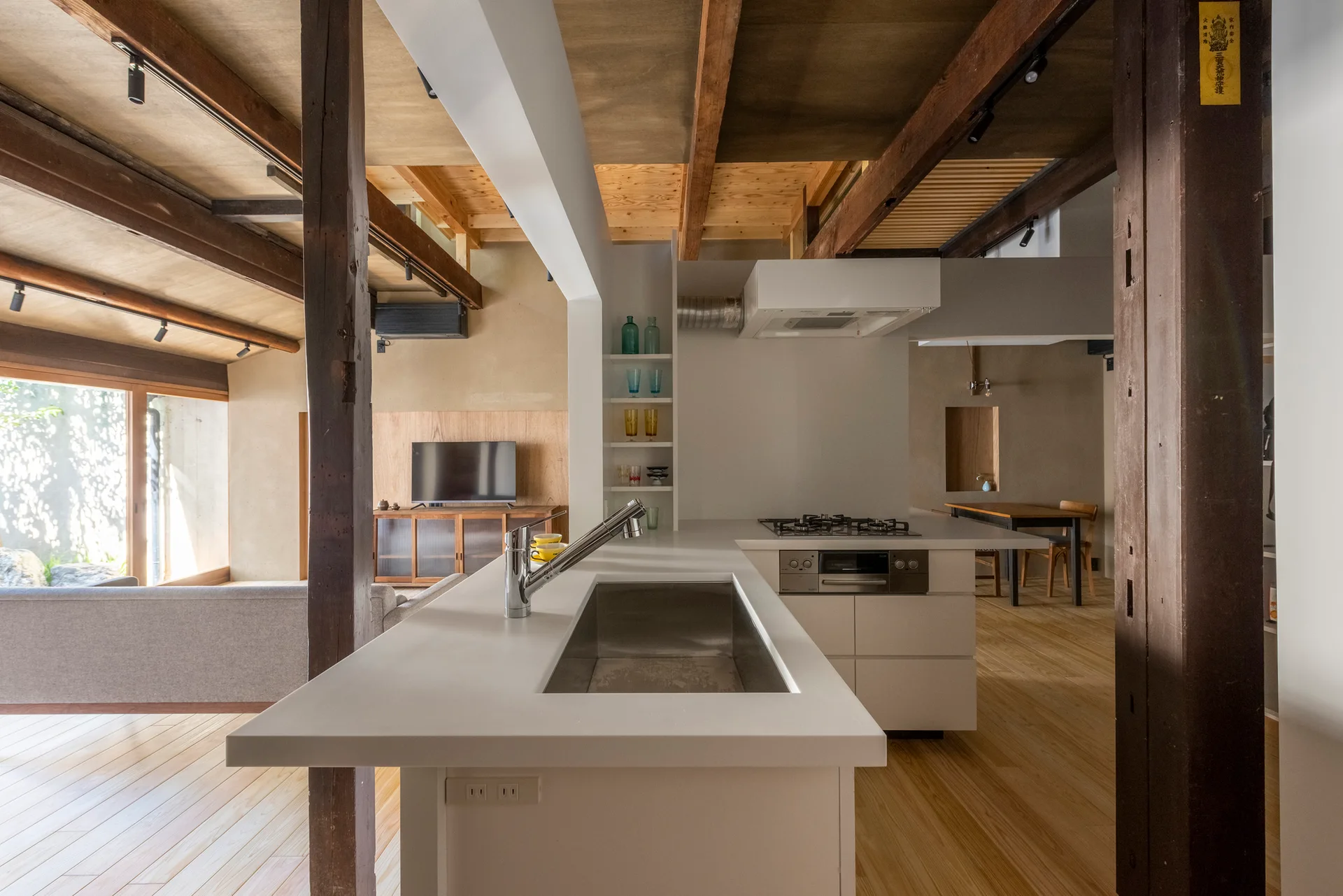 A minimalist white kitchen island with a metal sink sits beneath exposed dark wood ceiling beams and rustic timber columns in an open-plan home.