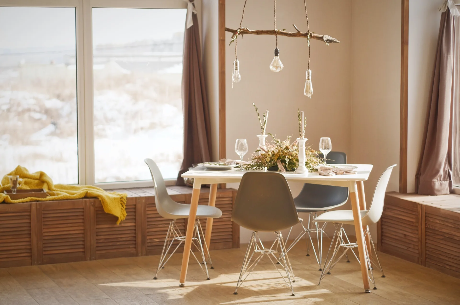 A bright dining room features a white table with mid-century style chairs under a rustic branch light next to wood window benches.