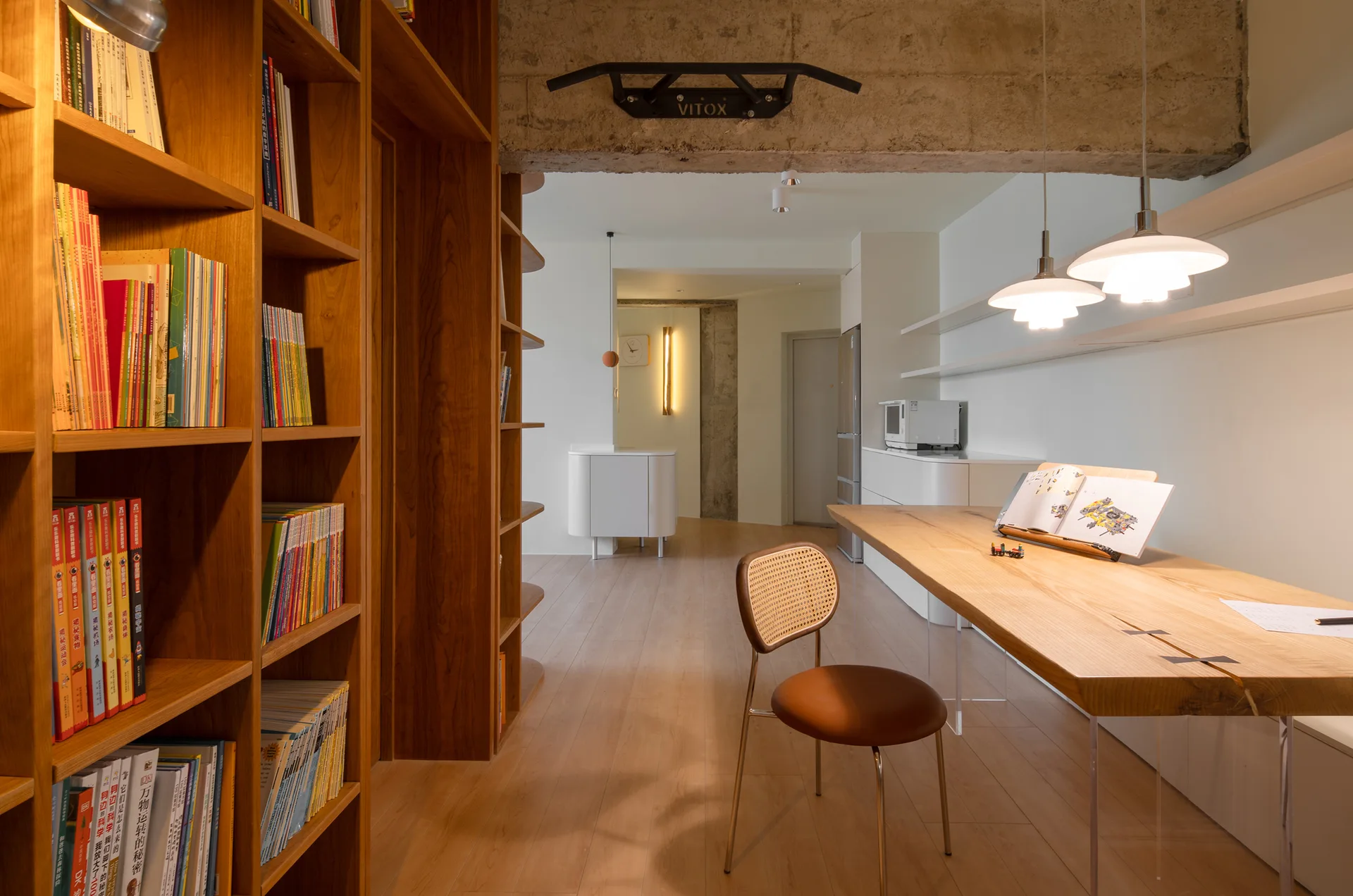 A study room featuring a large wooden bookcase, a natural wood desk with clear acrylic legs, and a cane chair under an exposed concrete beam.