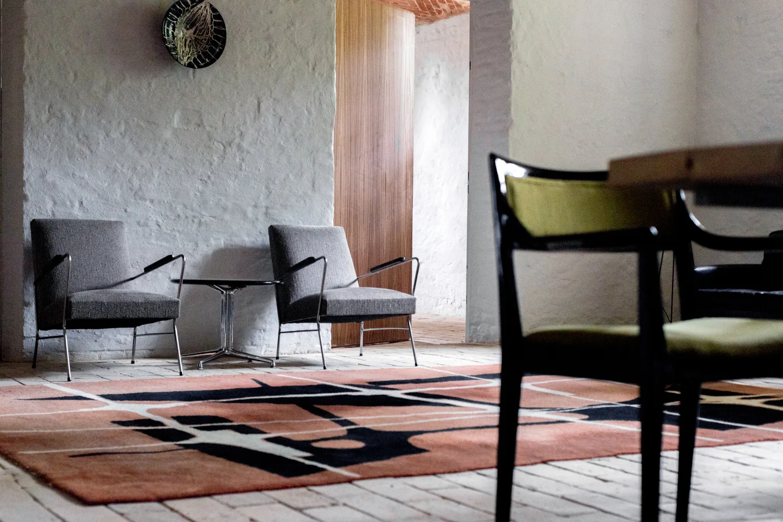 Two gray armchairs and a small side table sit on a geometric terracotta rug against textured white plaster walls.