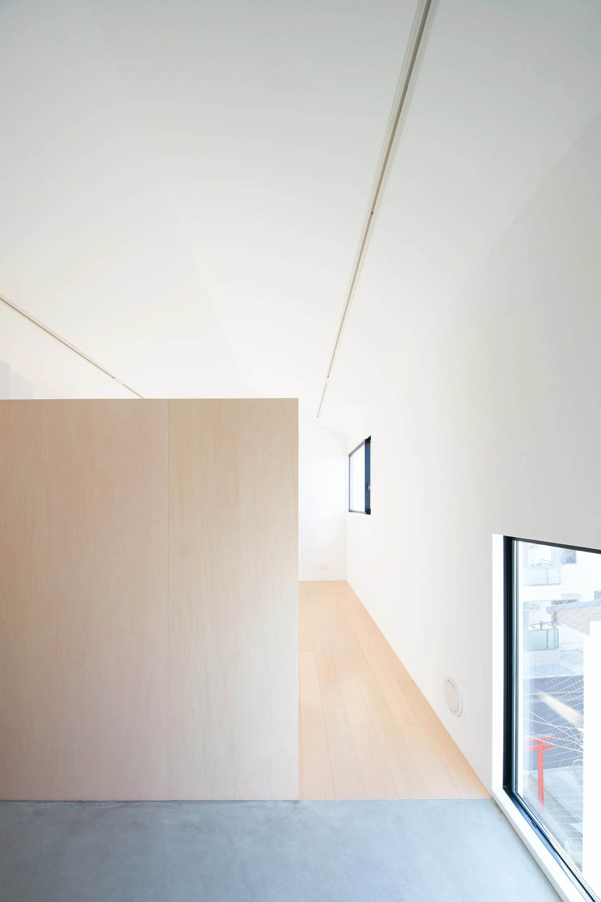 A minimalist room featuring a large plywood partition wall dividing light wood flooring and a smooth concrete floor near a window.