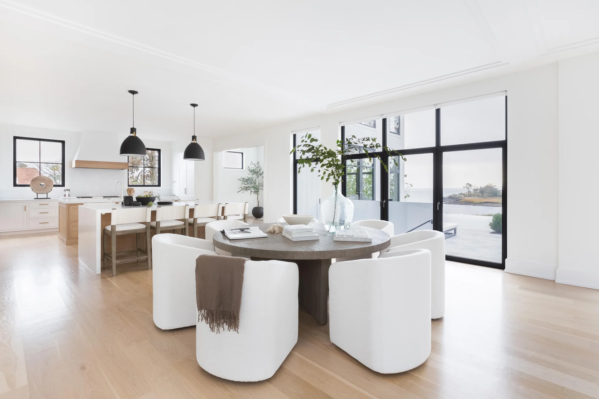 A modern dining room featuring a round wooden table, white upholstered barrel chairs, and large black-framed sliding glass doors with a waterfront view.