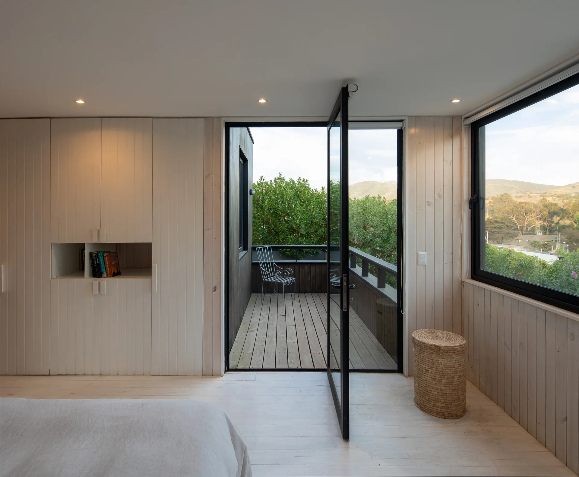 A minimalist bedroom featuring off-white vertical wood paneling, a built-in wardrobe, and a large open black-framed glass door leading to a balcony.