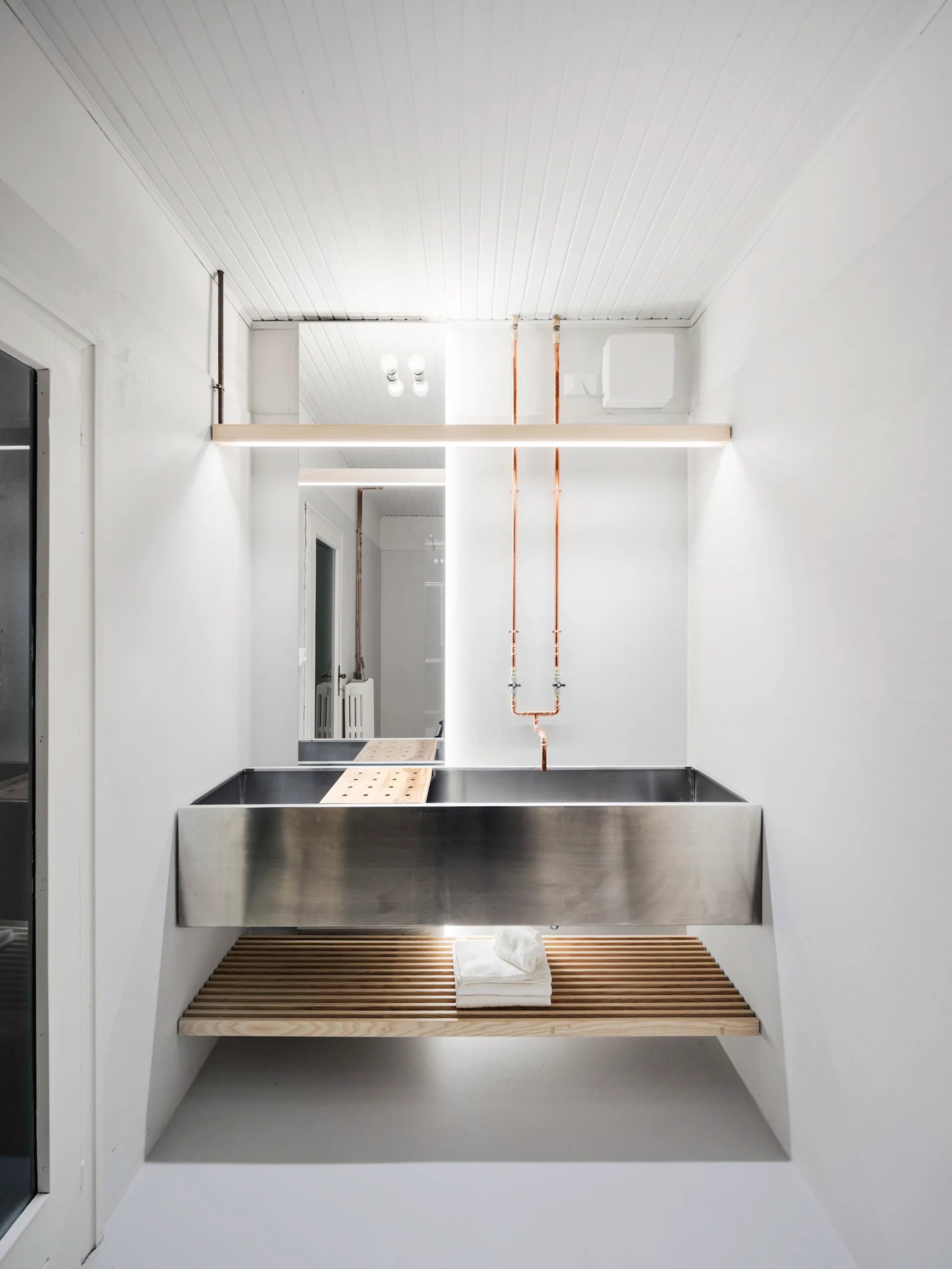 A minimalist bathroom features a wide stainless steel trough sink, exposed vertical copper plumbing, a wooden slatted lower shelf, and a tall mirror.
