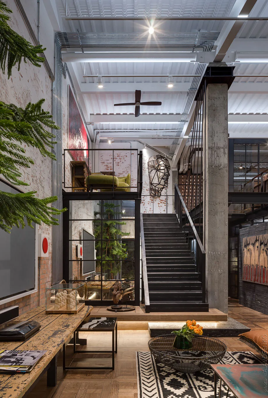 An industrial loft living space featuring a black metal staircase, a mezzanine level, exposed brick walls, and a reclaimed wood desk.