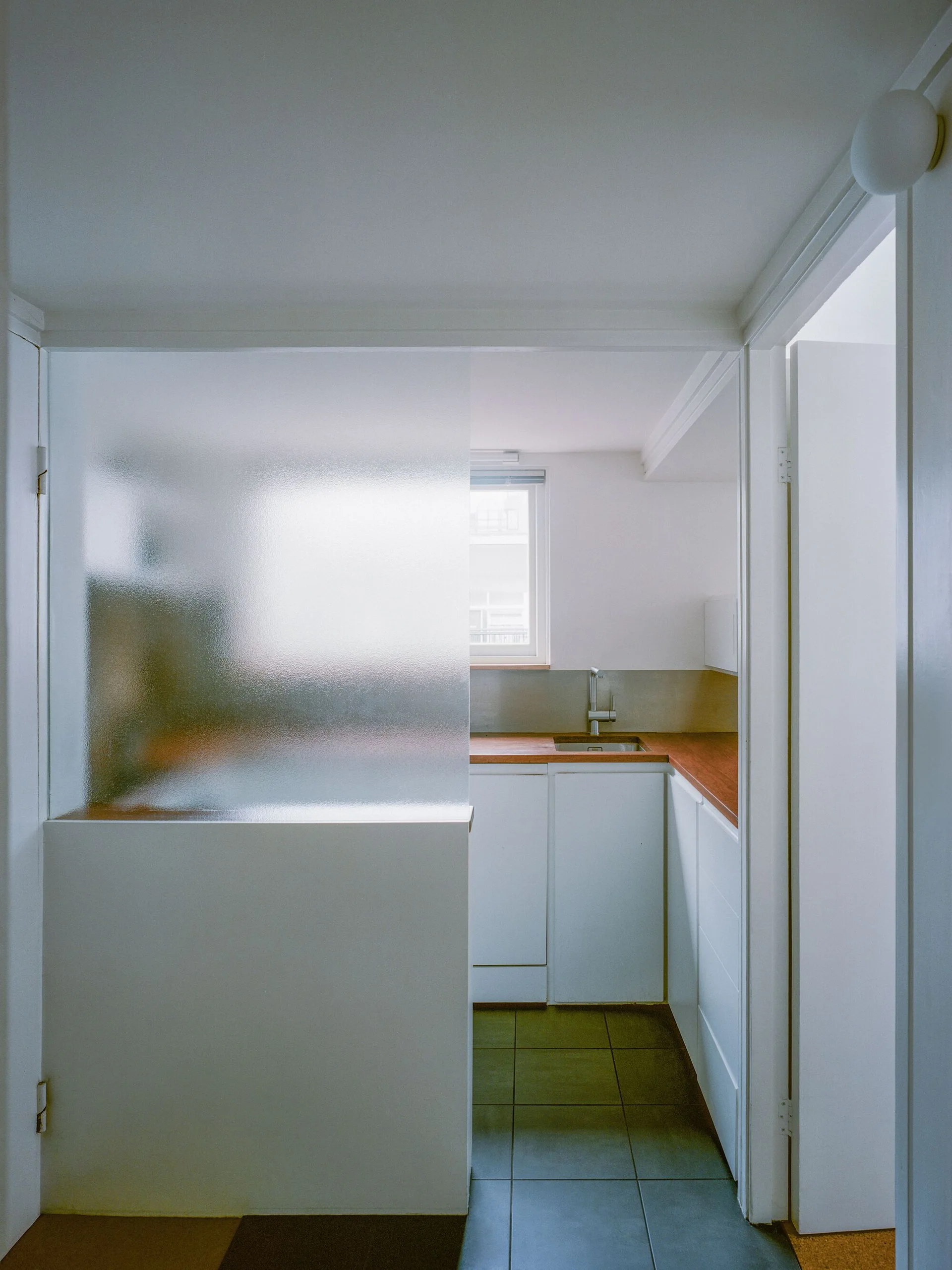 A minimalist kitchen seen through a doorway, featuring white cabinetry, warm wood countertops, grey tile flooring, and a frosted glass partition.