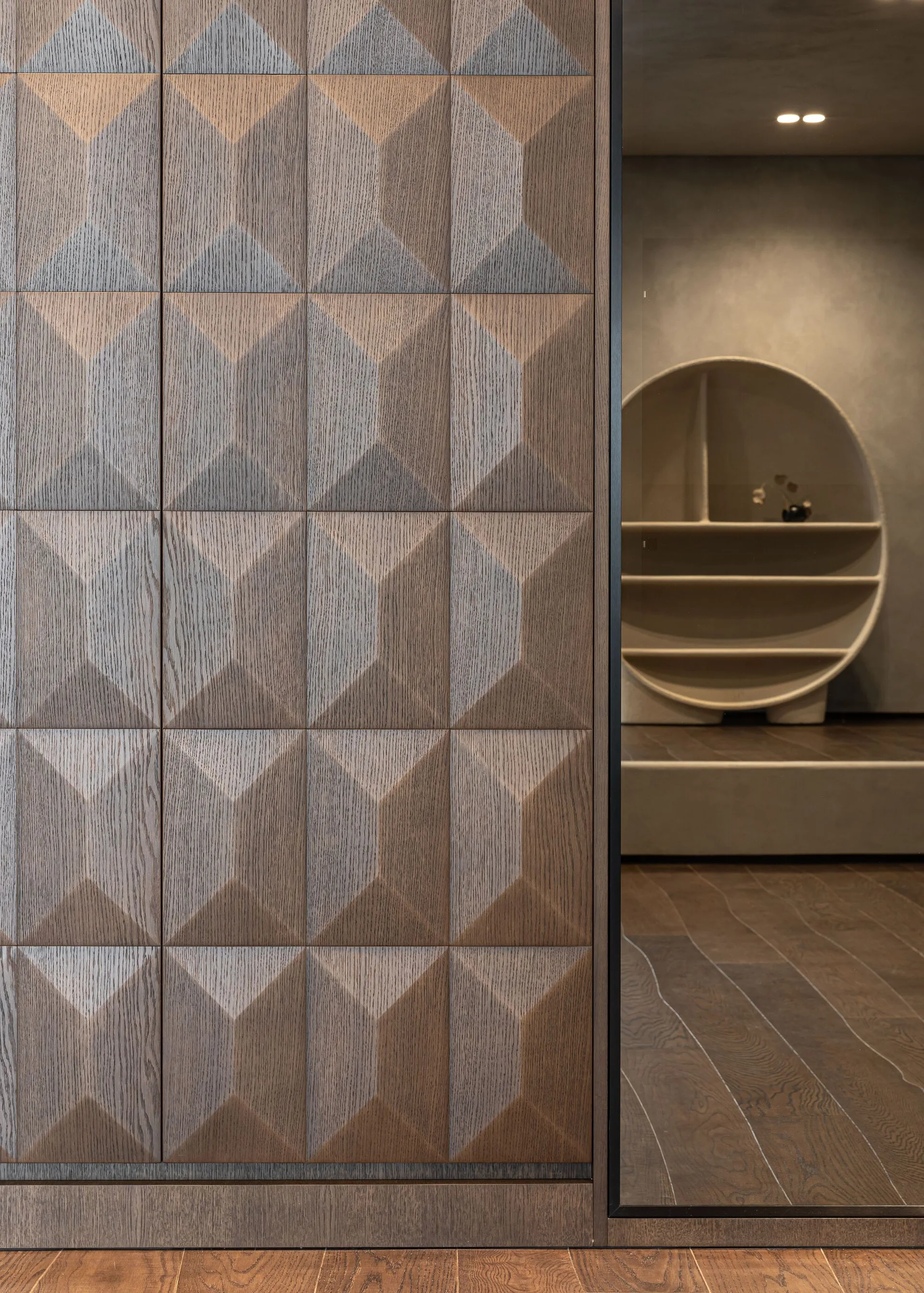 Faceted brown wood wall panels with a geometric pattern stand beside a doorway revealing a circular built-in shelf in a grey plastered wall.
