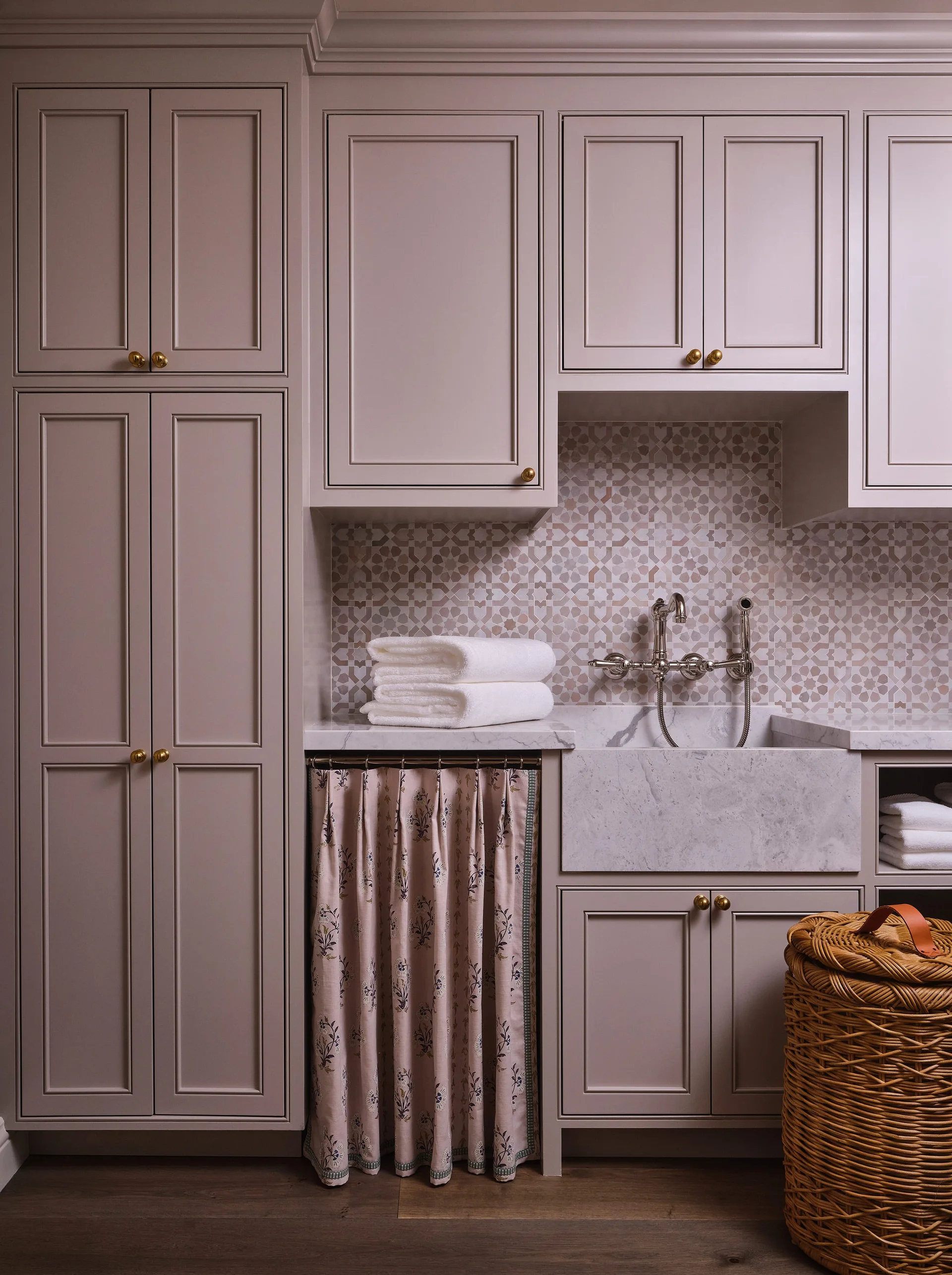A utility sink area features mauve shaker cabinets, a stone farmhouse sink with a wall-mounted faucet, and a floral fabric curtain skirt.