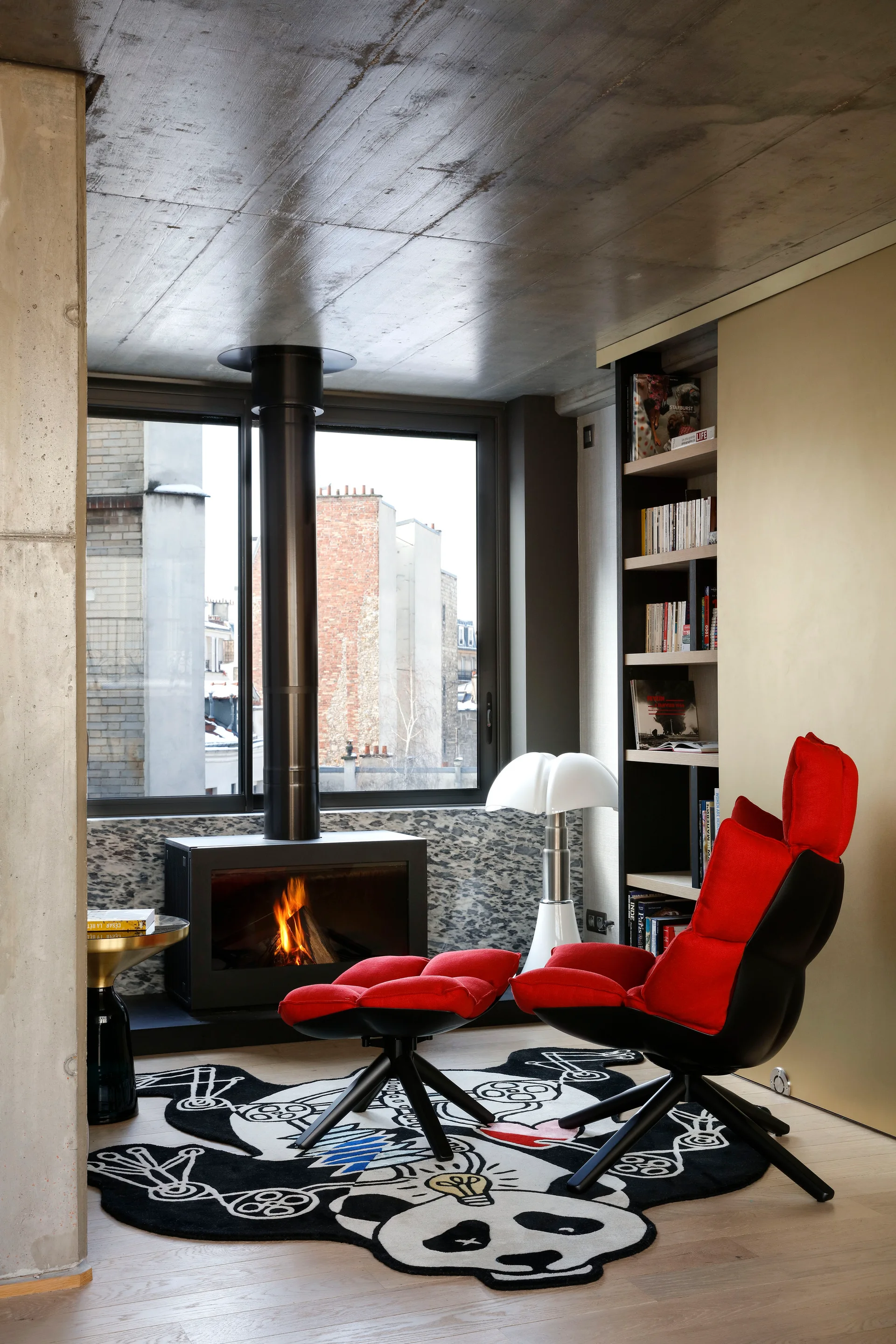 A modern lounge room featuring a red chair and ottoman set on a black and white graphic rug near a fireplace and a concrete ceiling.