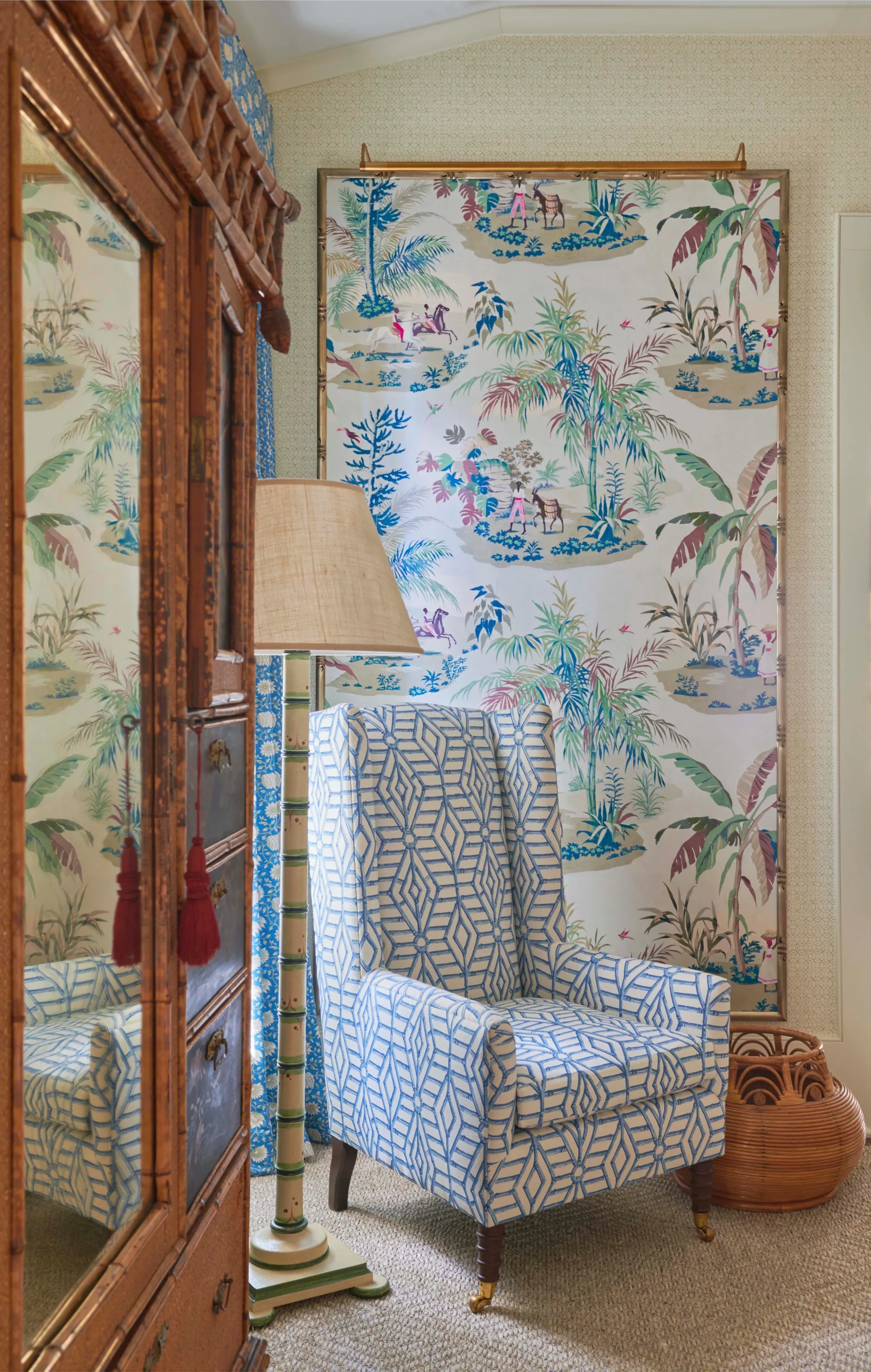 A traditional reading nook featuring a blue and white patterned wingback chair, a faux-bamboo wooden armoire, and a framed pictorial wallpaper panel.