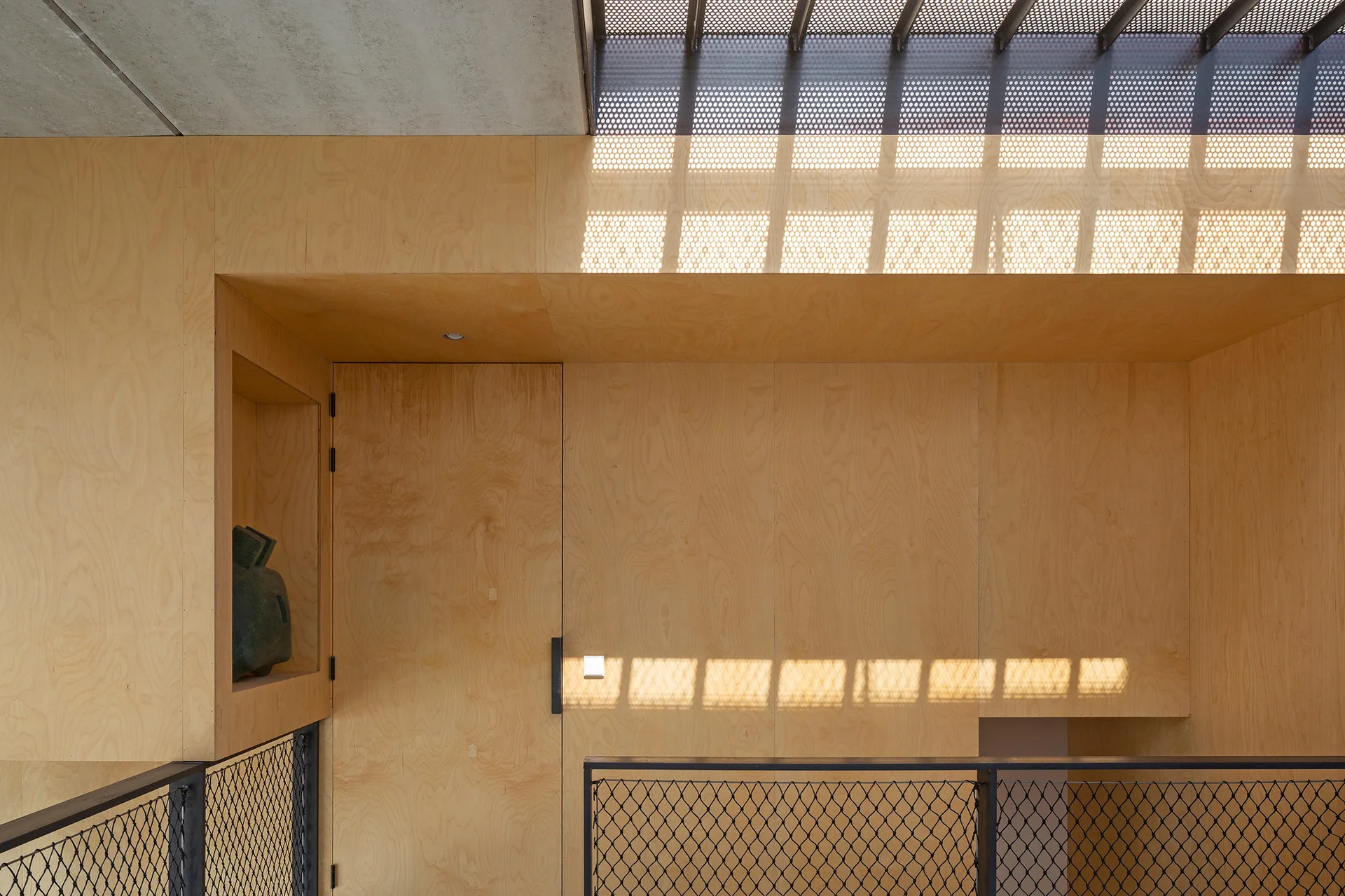 A modern staircase landing features light plywood wall paneling, a recessed display niche, black mesh railing, and a perforated metal ceiling skylight.
