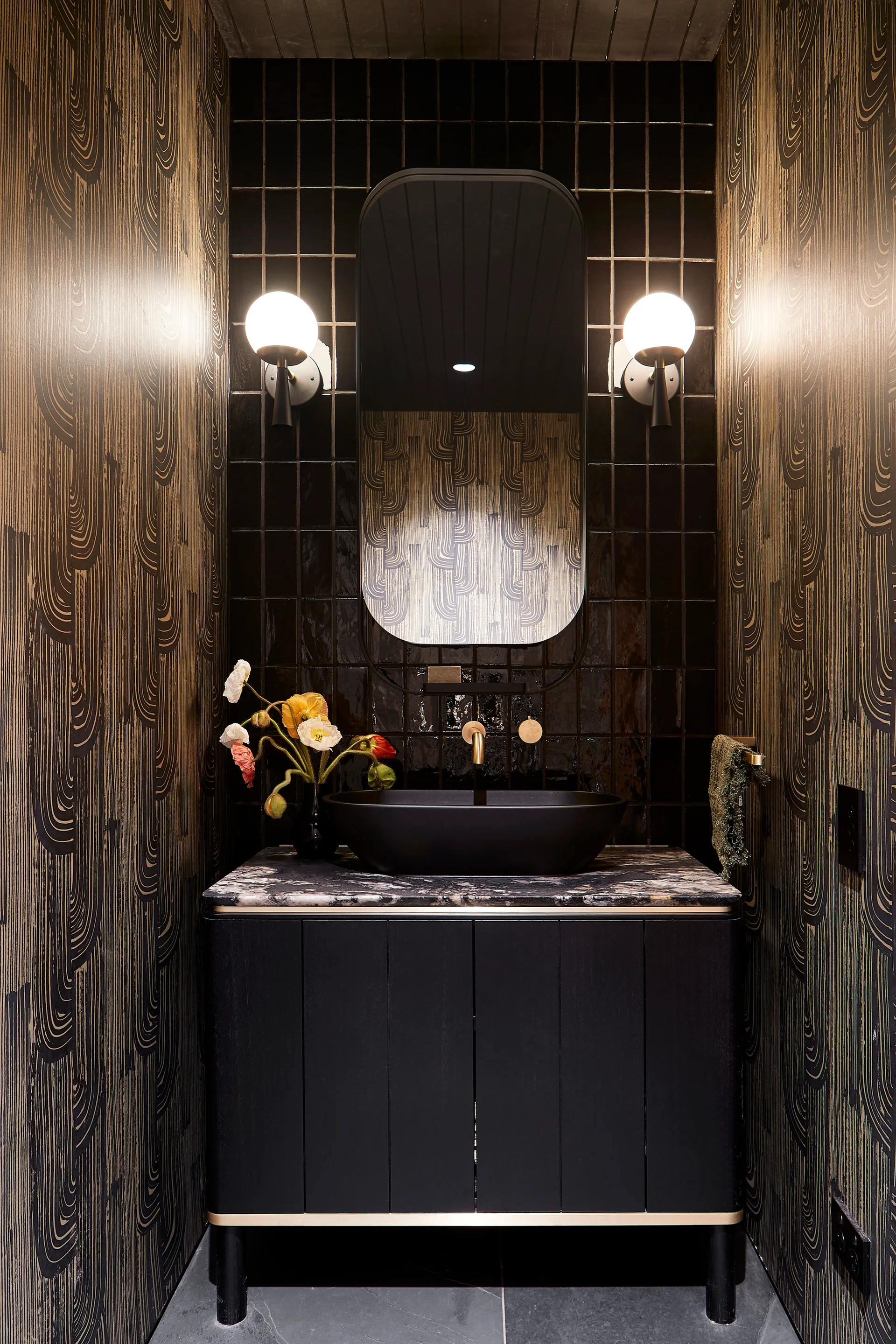 A dark powder room featuring a black vessel sink on a veined stone vanity, flanked by graphic patterned wallpaper and a glossy tile wall backsplash.
