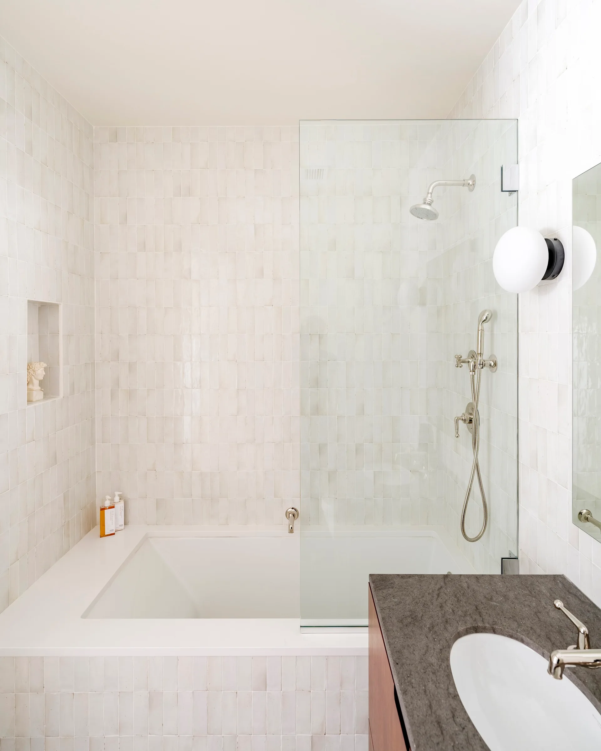 A modern bathroom features a white alcove bathtub, vertically stacked off-white wall tiles, a glass shower screen, and a dark stone vanity.