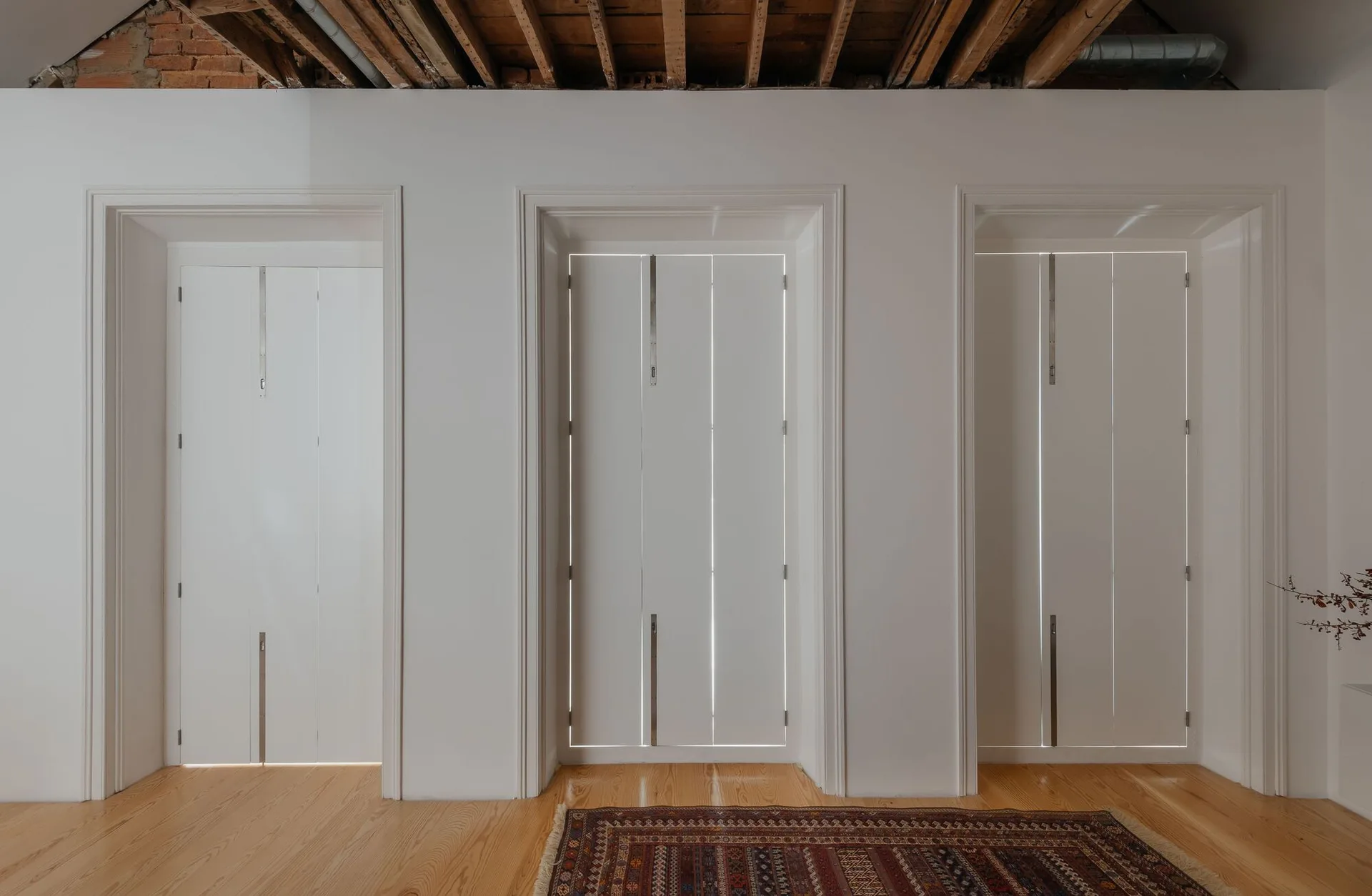 Three tall, white folding doors with silver hardware are set into a smooth white wall beneath an exposed wooden ceiling with visible joists.