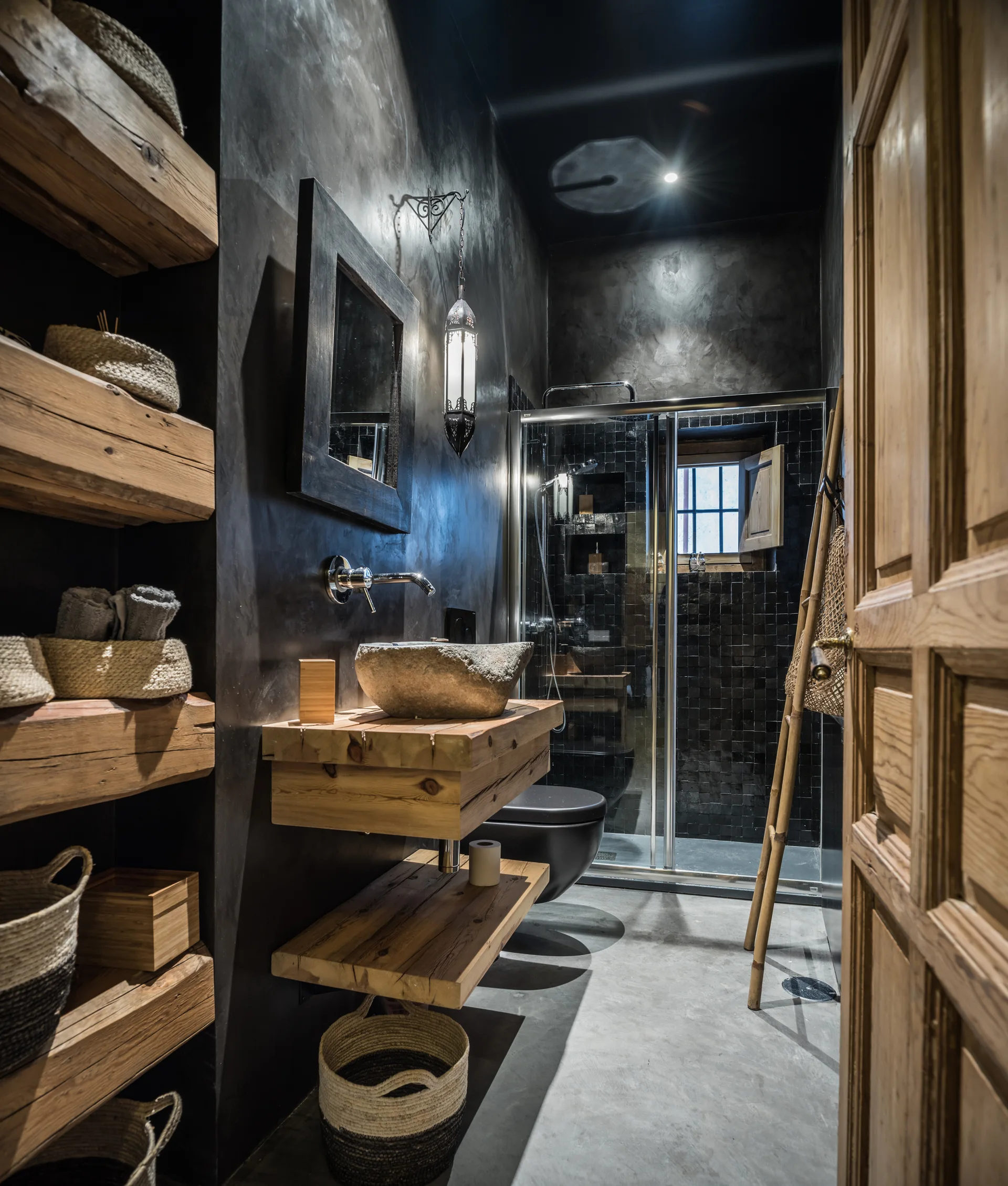 A rustic bathroom with dark textured plaster walls, thick floating wood shelves, a stone vessel sink on a timber vanity, and a glass-enclosed shower.
