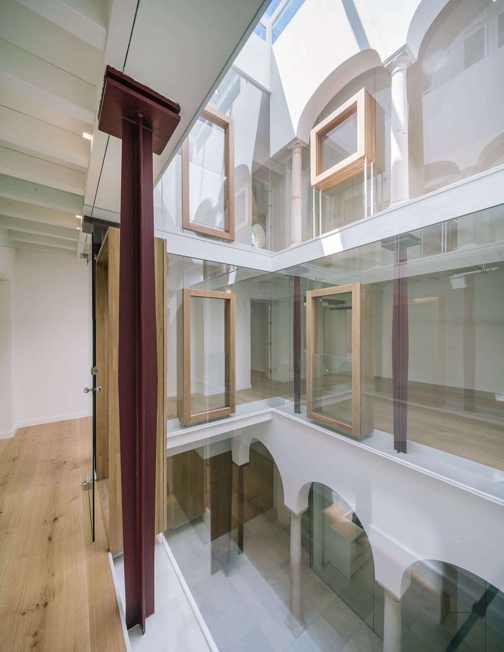 A multi-level interior atrium features floor-to-ceiling glass walls, arched plaster openings, stone columns, light wood floors, and a burgundy steel beam.