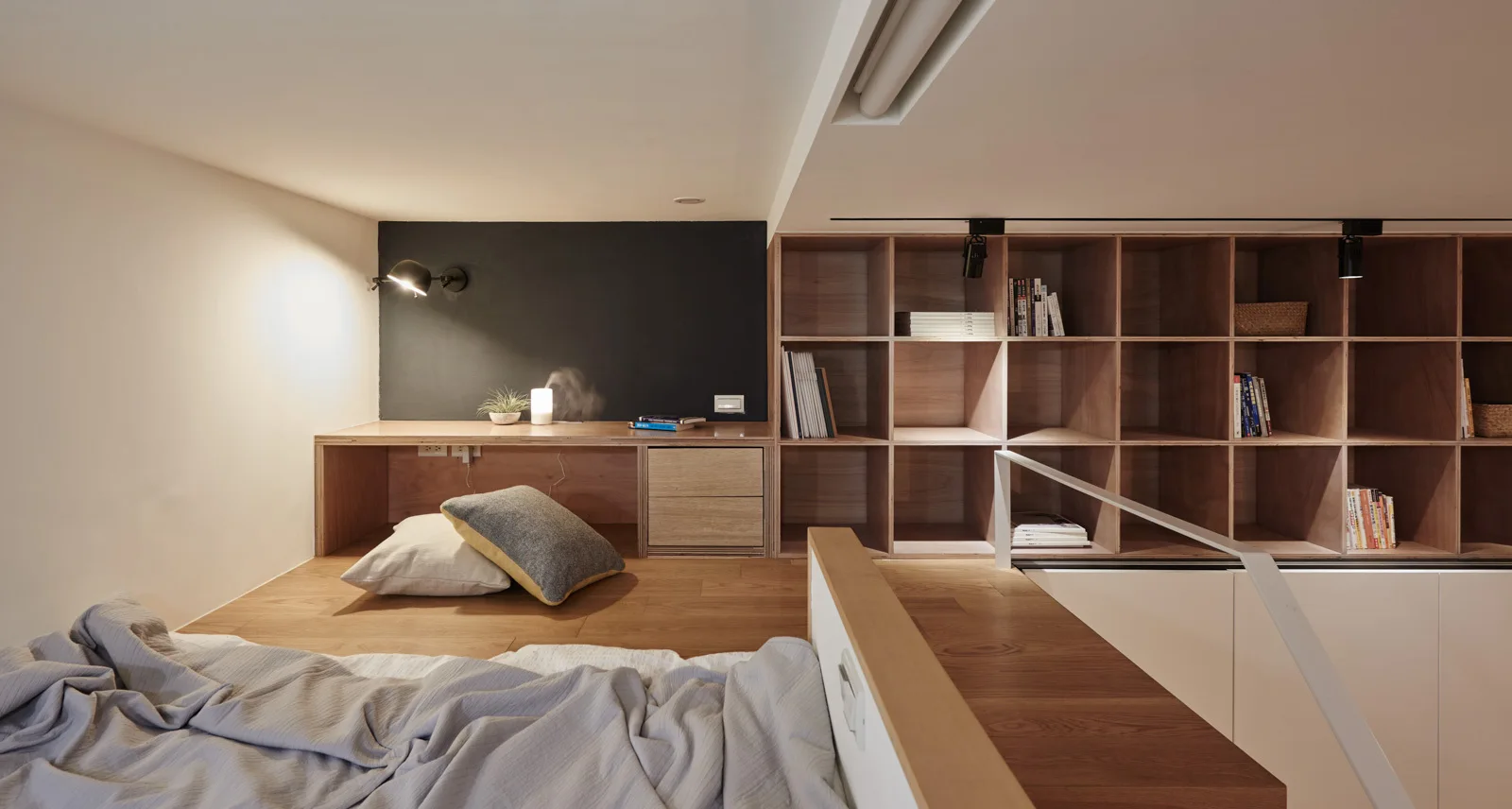 A minimalist loft bedroom featuring a wood platform with a built-in desk, black accent wall, and a large wooden grid shelving unit.