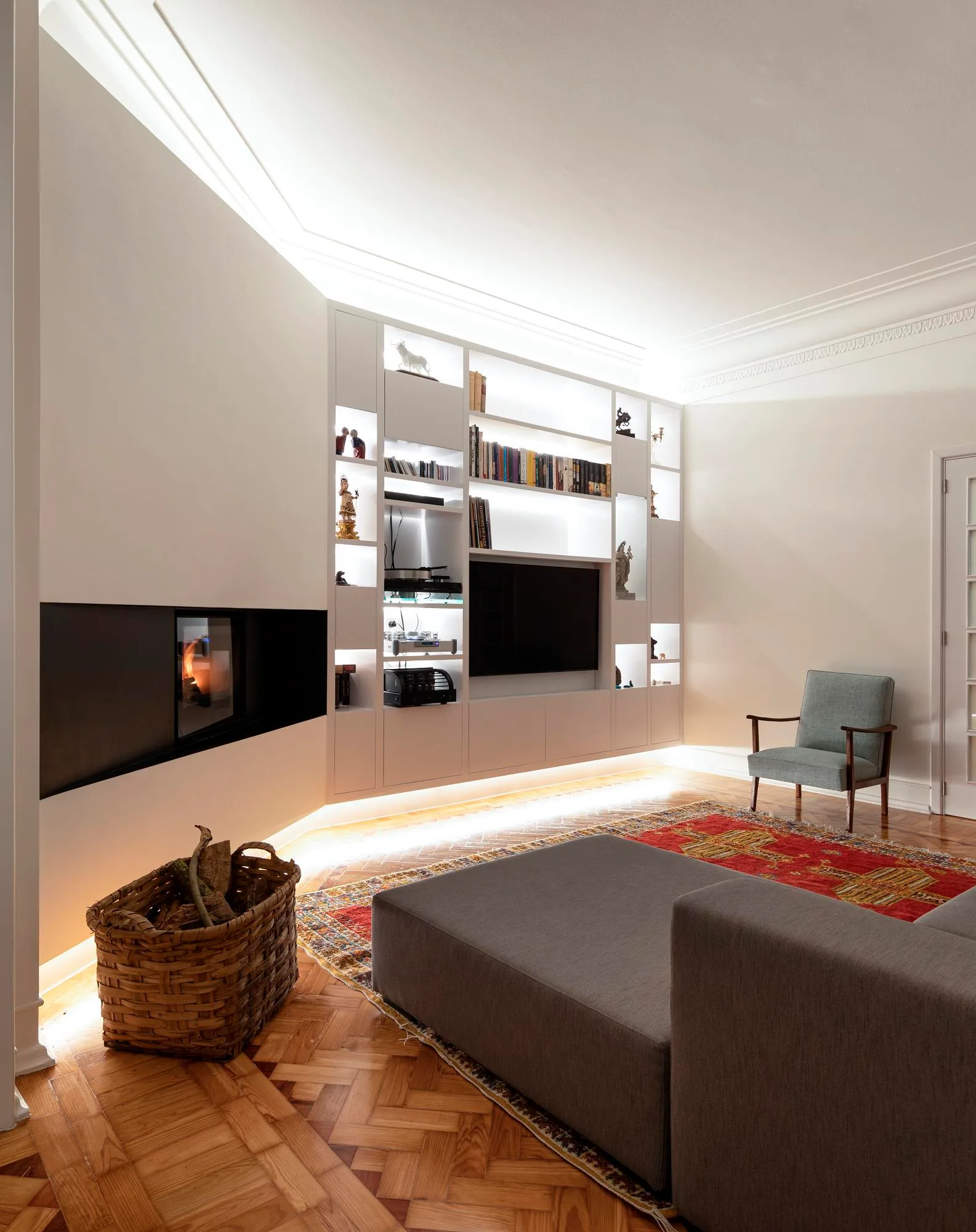 A living room features a white illuminated built-in shelving unit, a modern black fireplace, herringbone wood floors, and a red patterned rug.