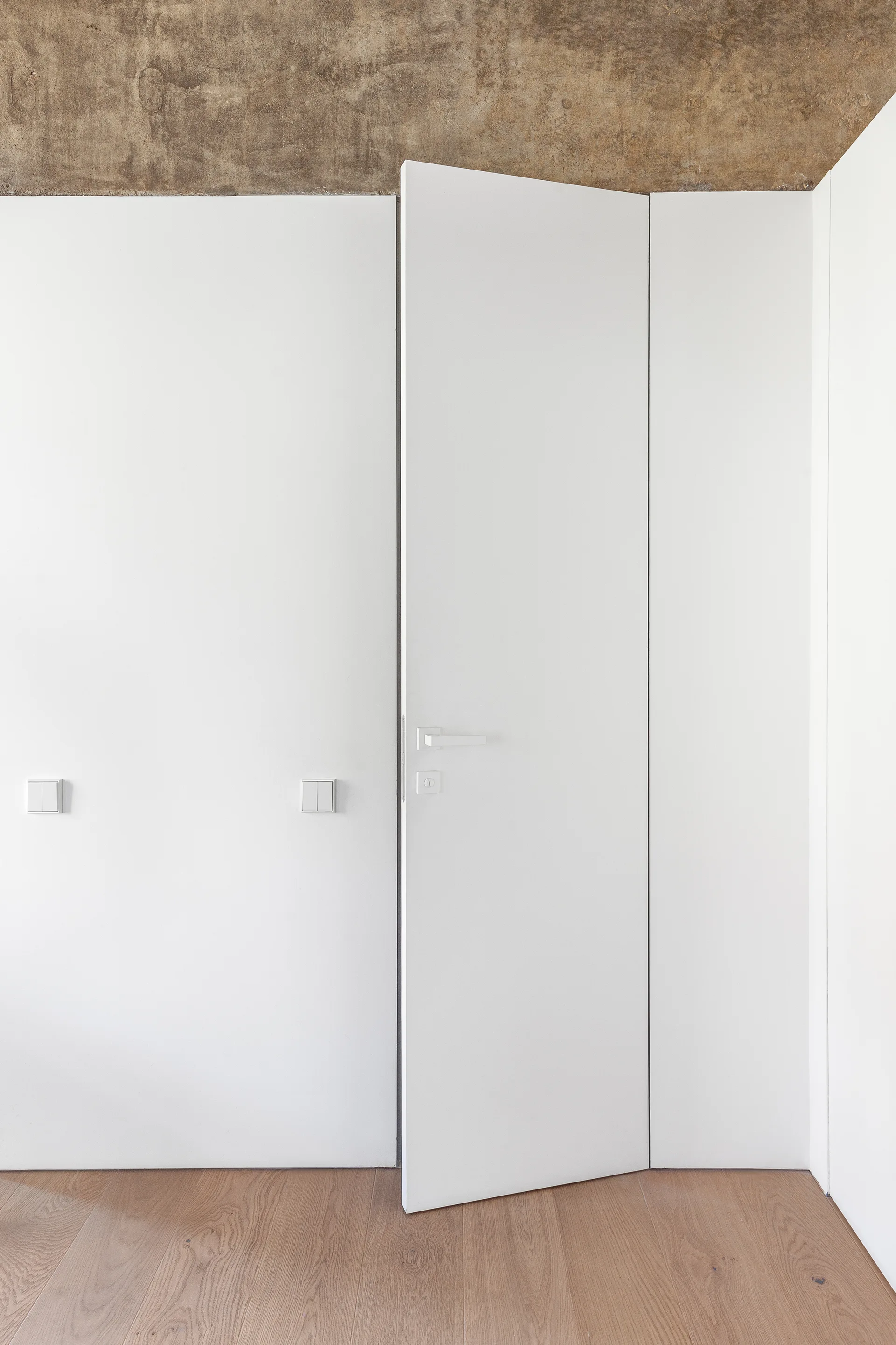 A partially open white flush-mount door stands against a smooth white wall beneath a raw concrete ceiling and above light wood flooring.