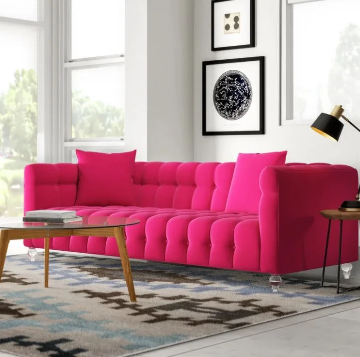 A vibrant hot pink tufted sofa with clear legs sits on a geometric patterned rug next to a glass coffee table in a bright living room.