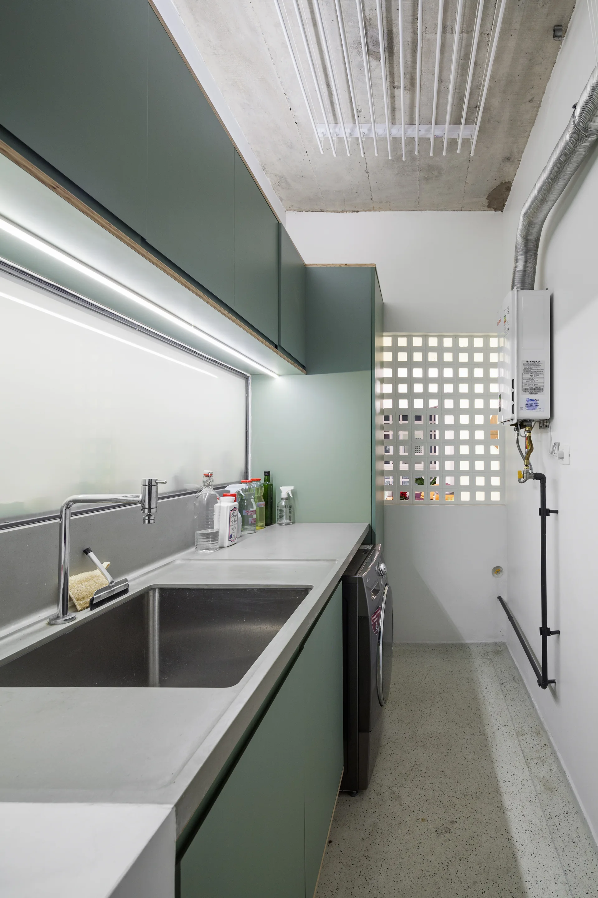 A minimalist laundry room features sage green cabinets, an under-cabinet light strip over a stainless steel sink, and a glass block window.