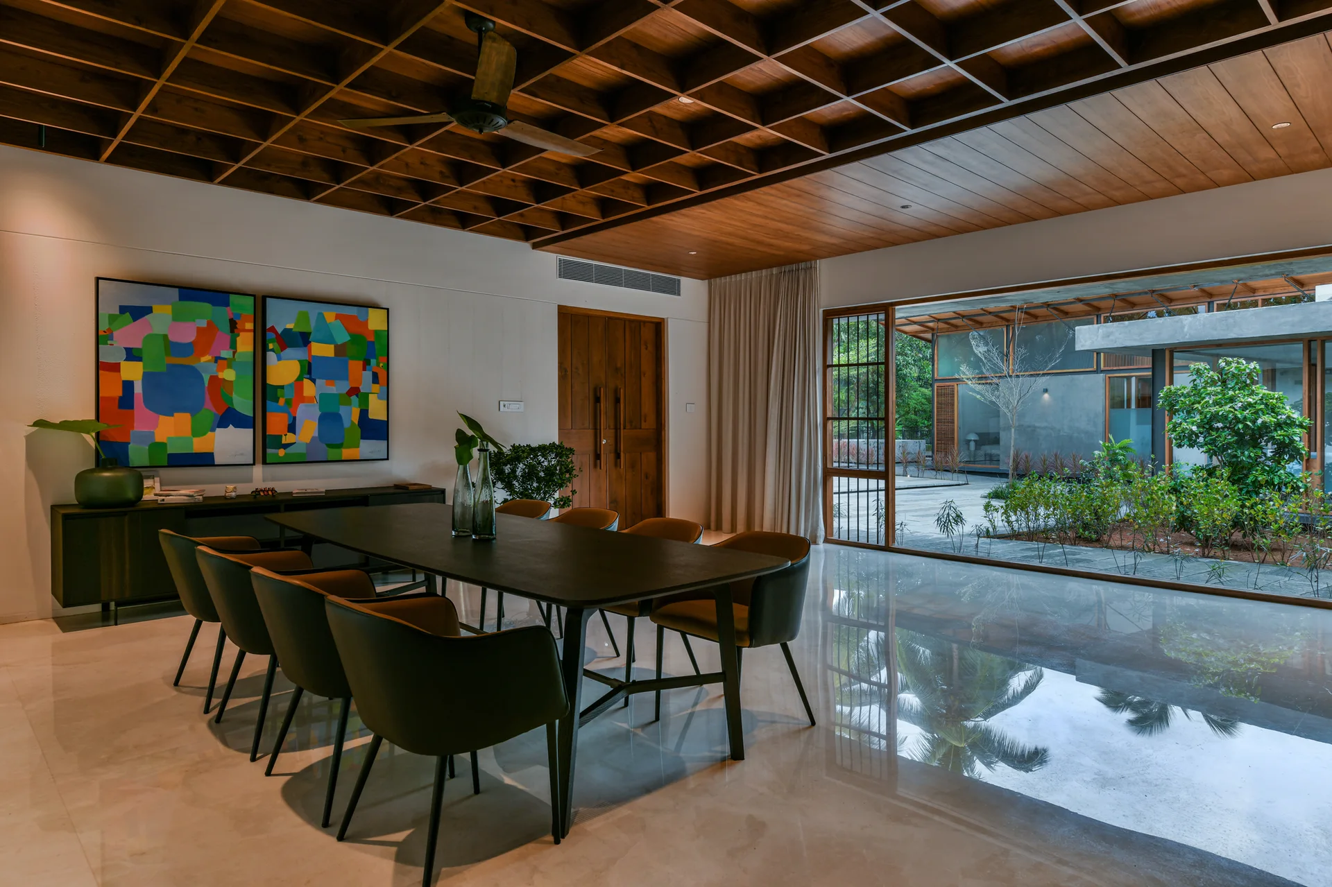 A modern dining room features a dark table, a wood coffered ceiling, and large glass doors opening to a lush green courtyard.