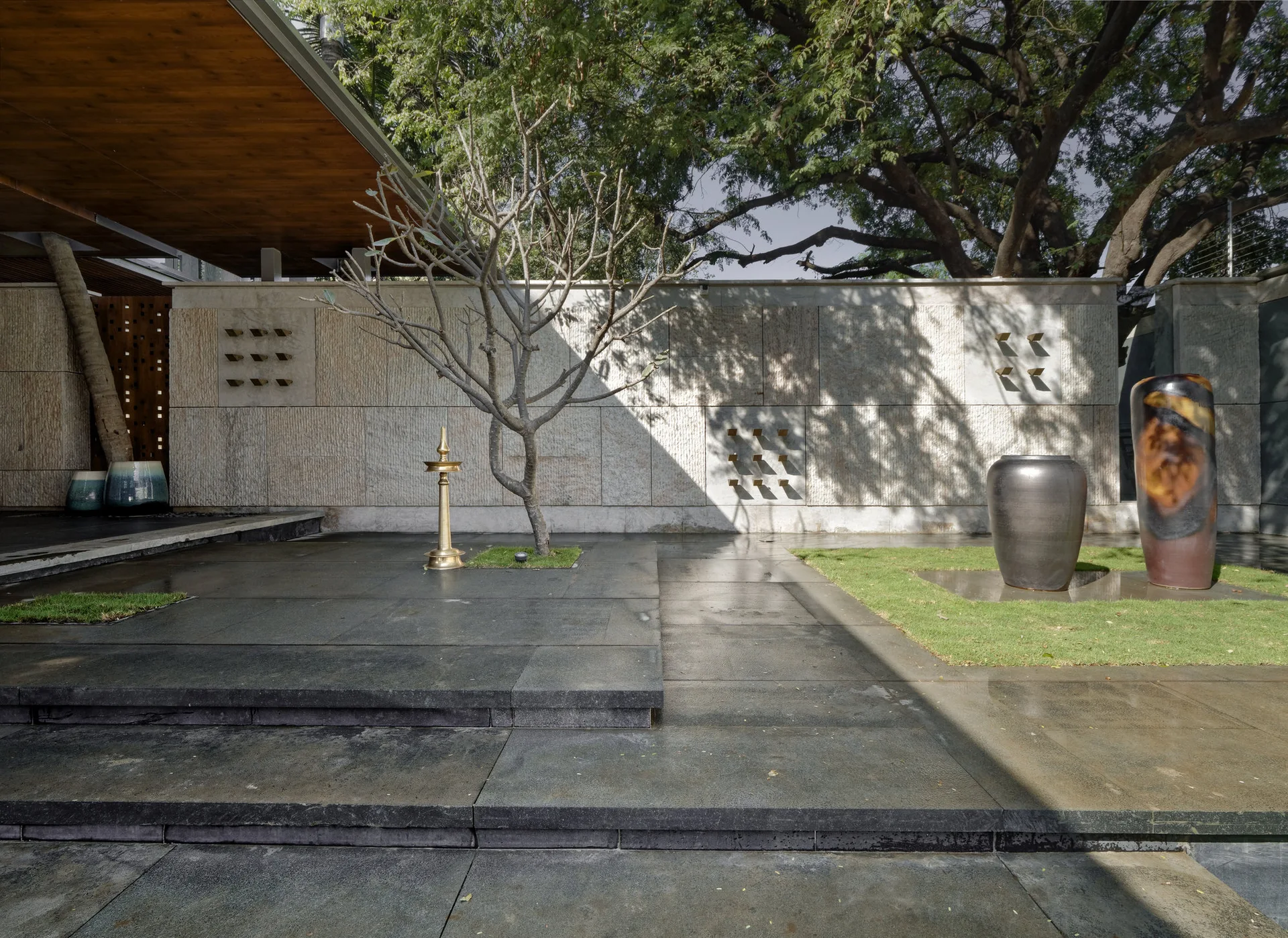 A minimalist outdoor patio with dark stone tile paving, a textured stone feature wall, a sculptural bare tree, and large ceramic urns.