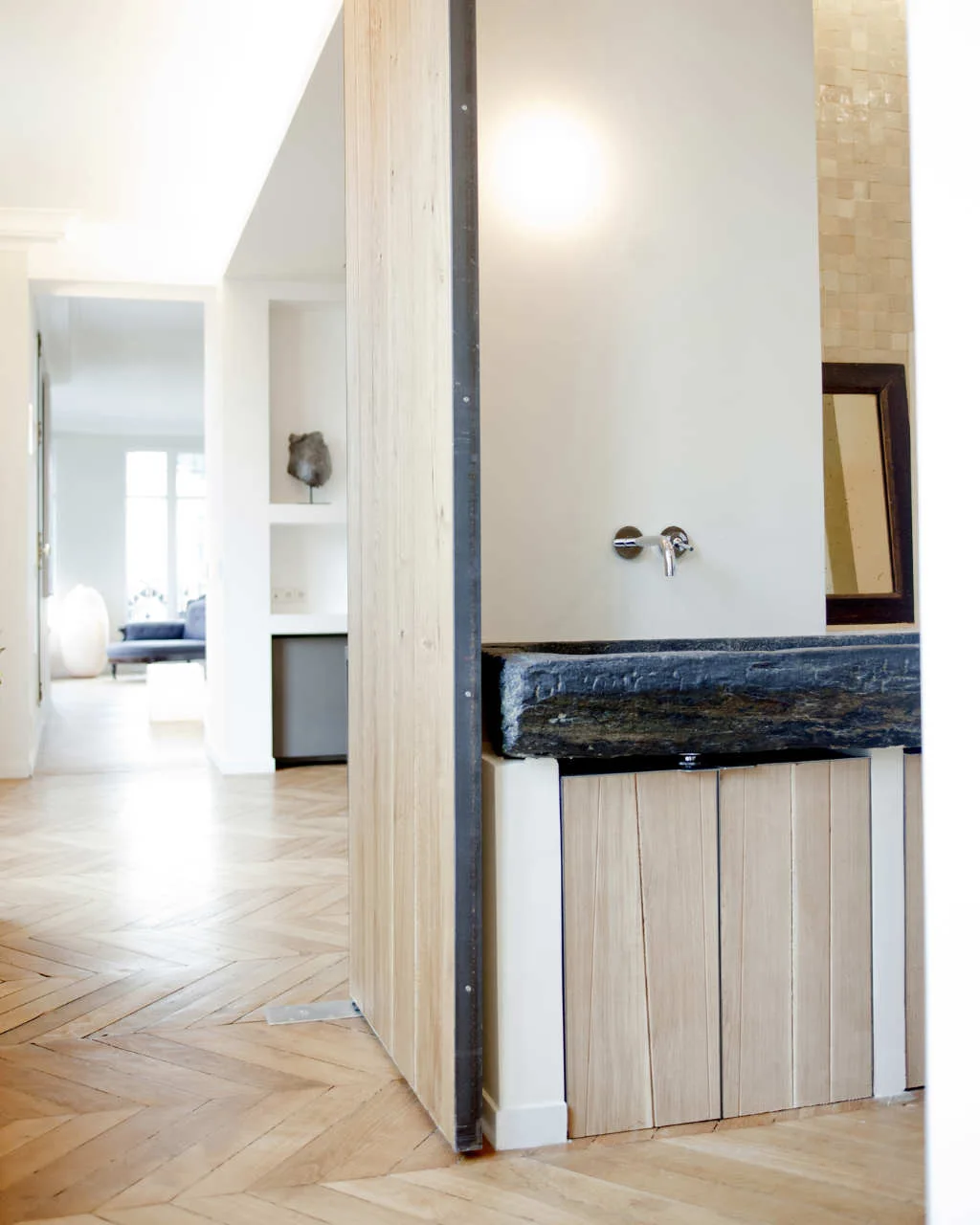 A dark stone trough basin sits on a light wood-paneled vanity beside a wall-mounted faucet and herringbone flooring.