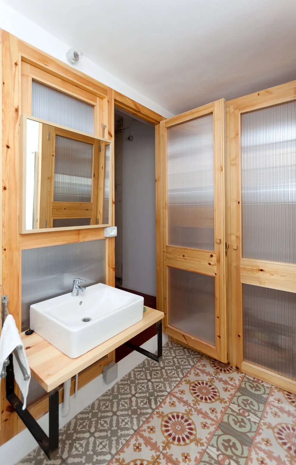 A modern bathroom vanity area features a white vessel sink on a wood shelf next to wooden doors with ribbed translucent panels and patterned floor tiles.