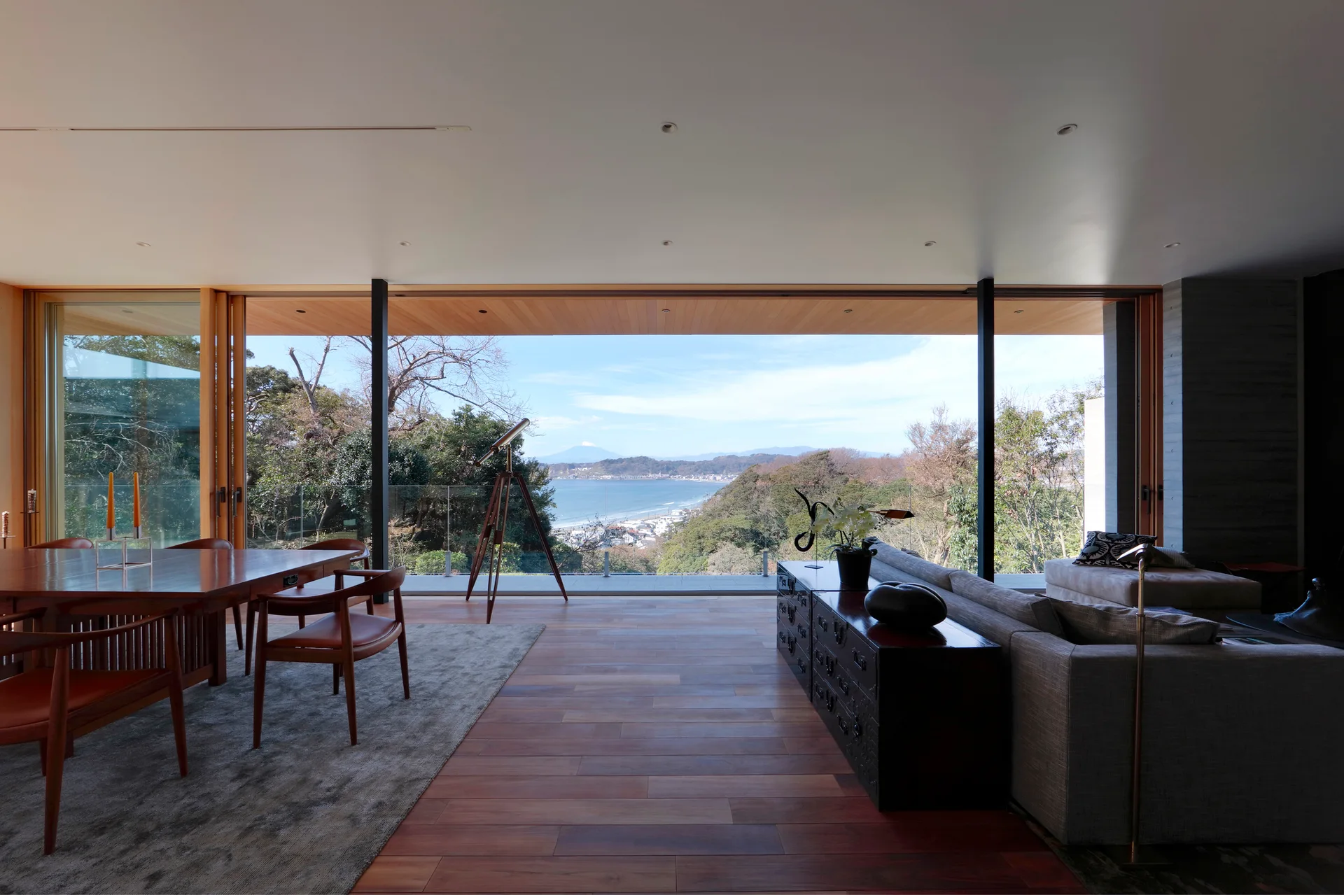 An open-concept living and dining room with a grey sectional, wood table, and floor-to-ceiling glass walls showcasing a coastal ocean view.