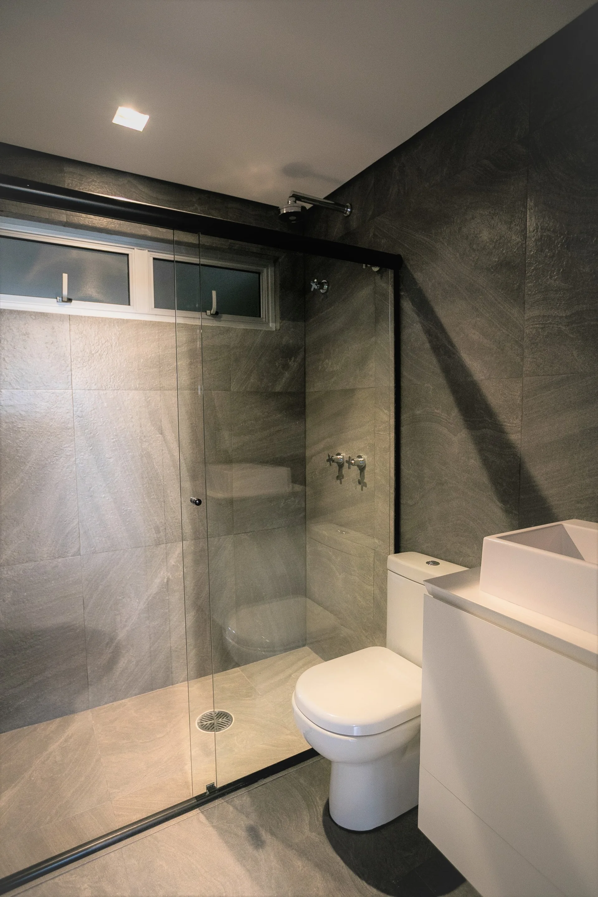 A modern bathroom features charcoal grey stone wall tiles, a glass shower enclosure with a transom window, a white toilet, and a white vanity.