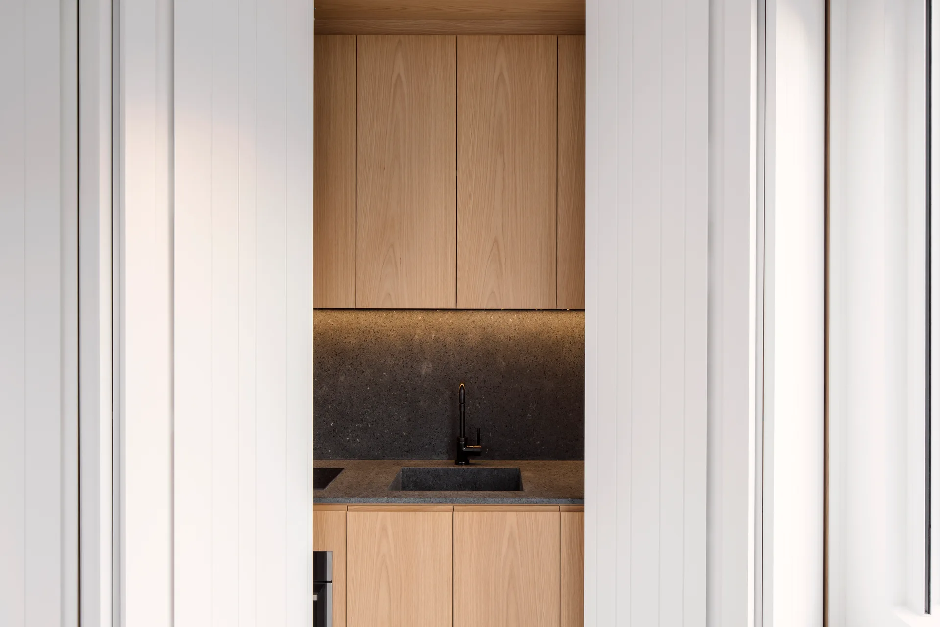 A modern kitchen niche with light wood cabinets, a dark stone countertop, and a matte black faucet framed by white paneled walls.