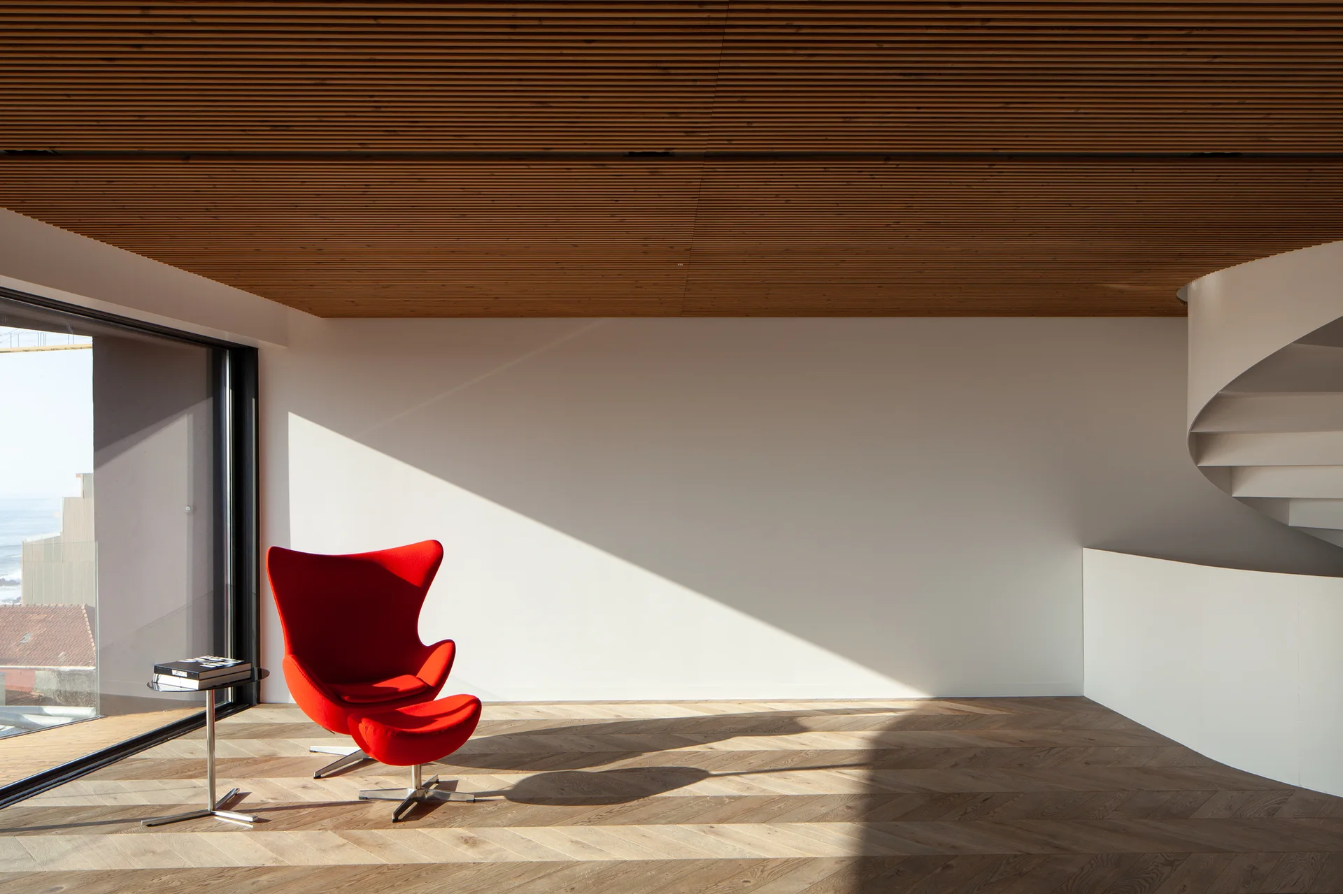 A modern room features a red egg chair and matching ottoman on herringbone wood floors beneath a slatted timber ceiling.
