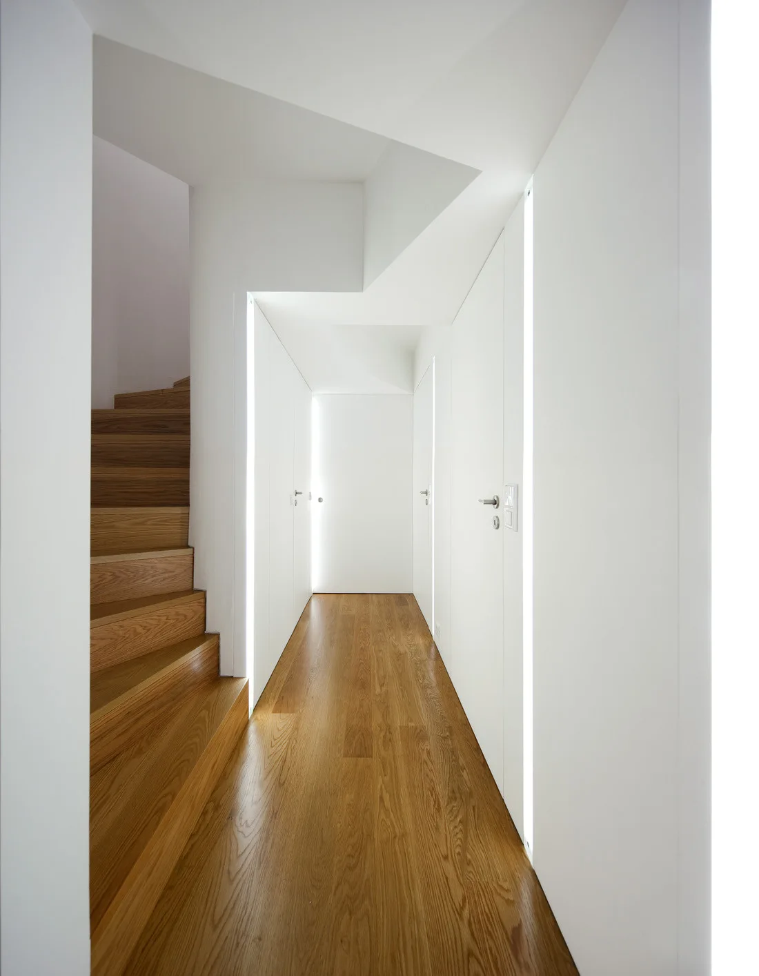 A minimalist white hallway features natural wood flooring, a matching wooden staircase on the left, and doors framed by vertical strip lights.